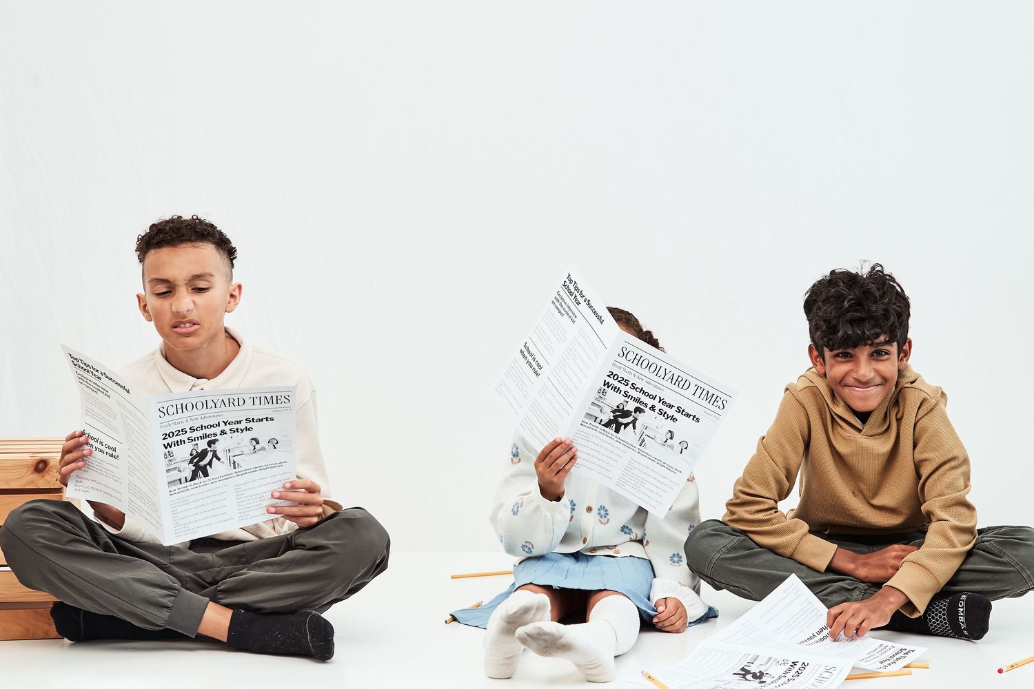 Three children sitting cross-legged on the floor reading newspapers in a minimalist white setting.
