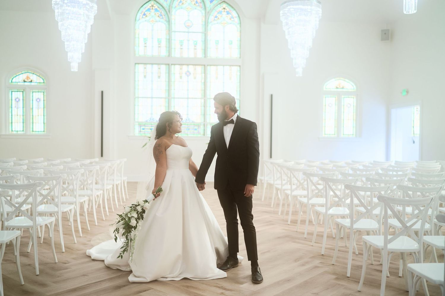 Dreamy wedding chapel scene with rows of white chairs and large arched stained glass windows.
