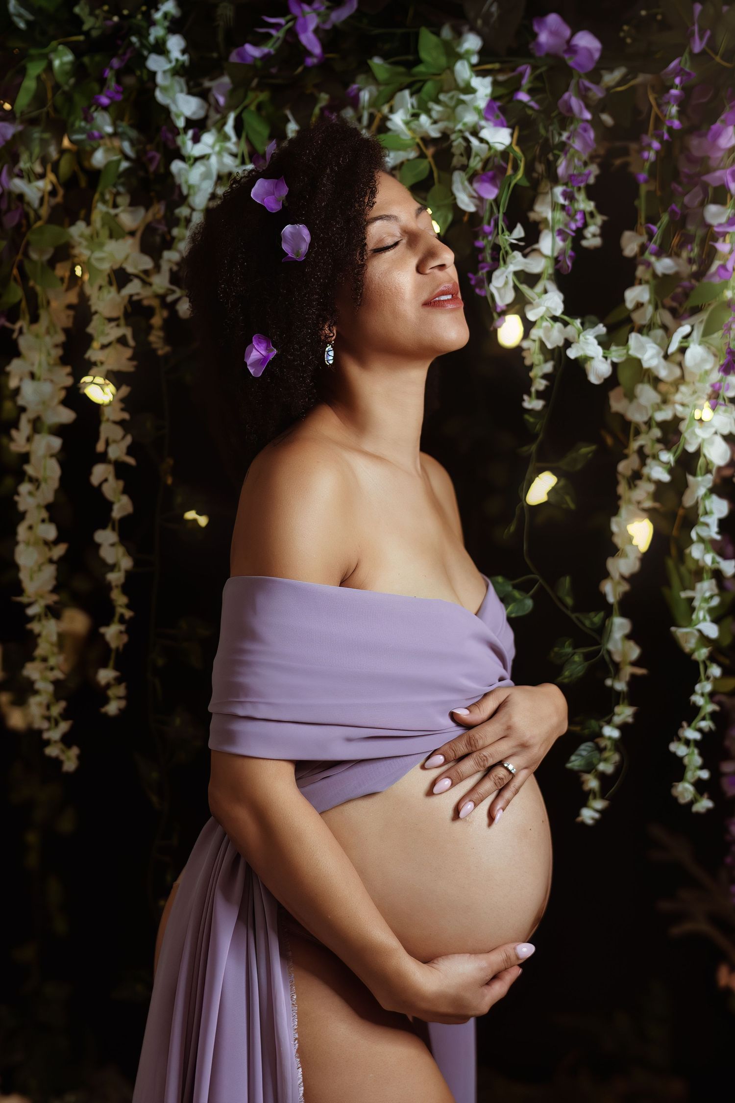 A maternity photoshoot with a flowing lavender dress against a backdrop of hanging floral decorations.