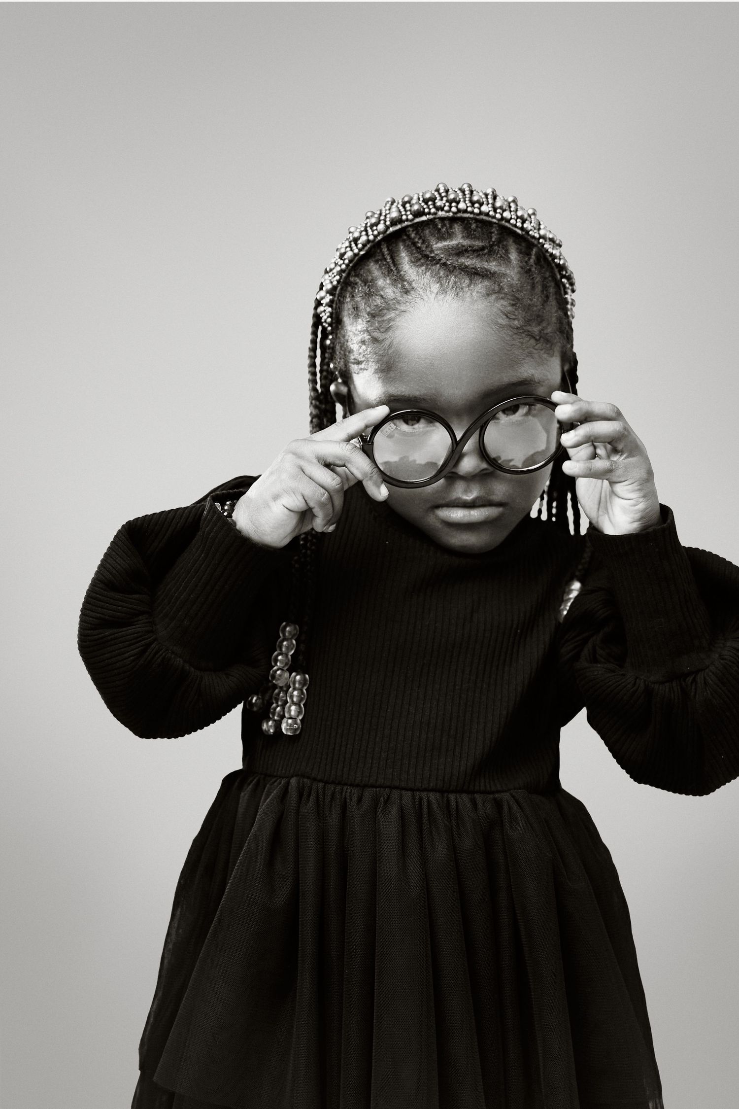 A child in a black dress adjusts heart-shaped glasses while wearing braids and a decorative headband in black and white.