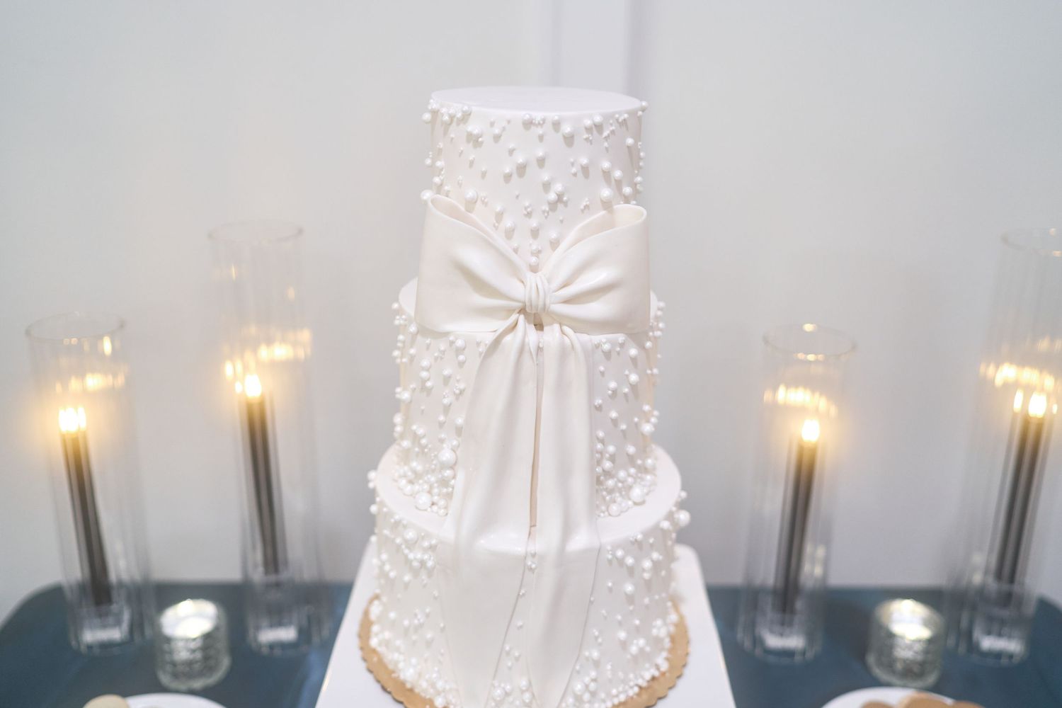 An elegant white wedding cake with pearl details and a large bow decoration, surrounded by glowing candles.