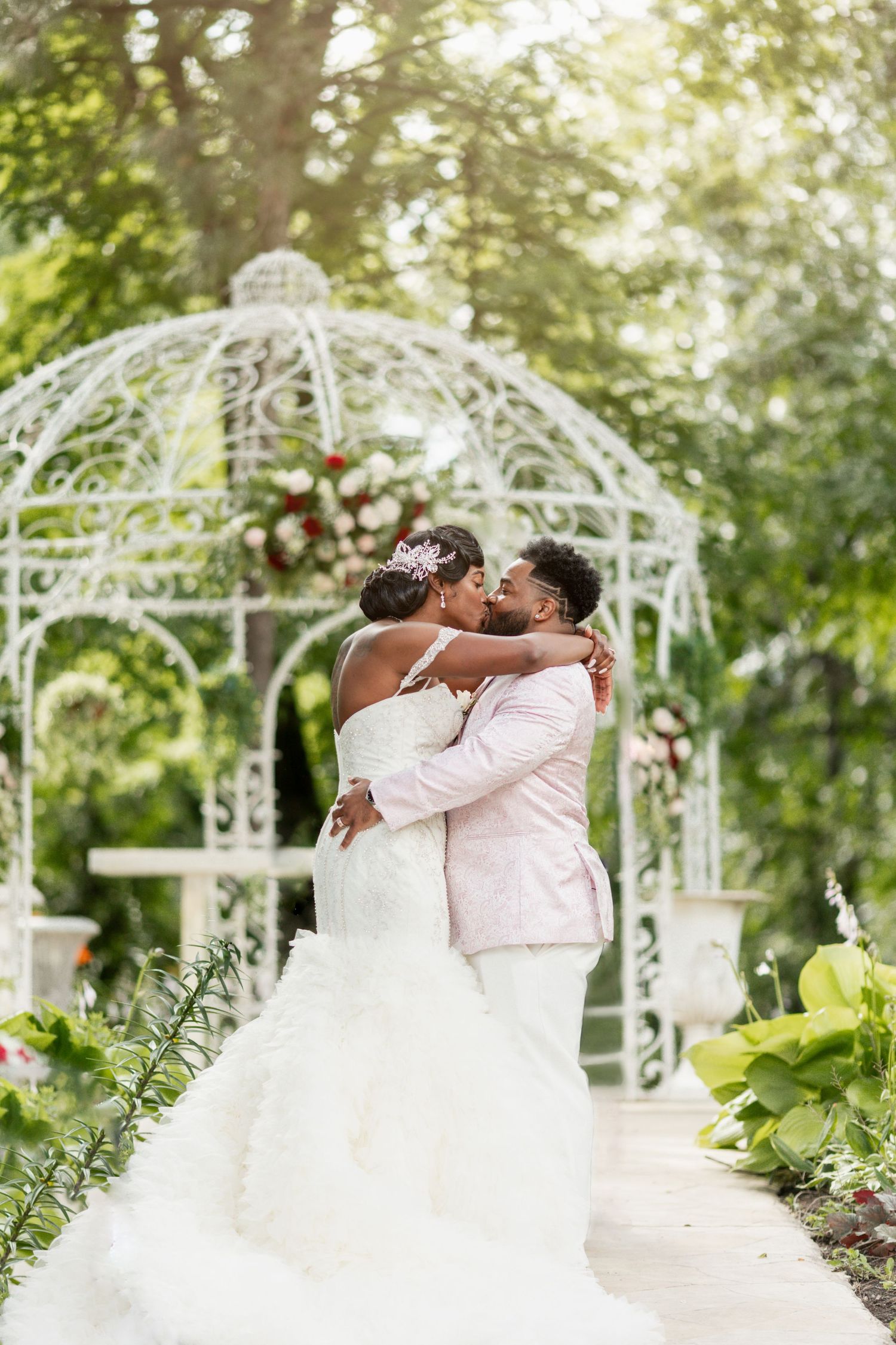 Newlyweds share first kiss under ornate white garden gazebo.