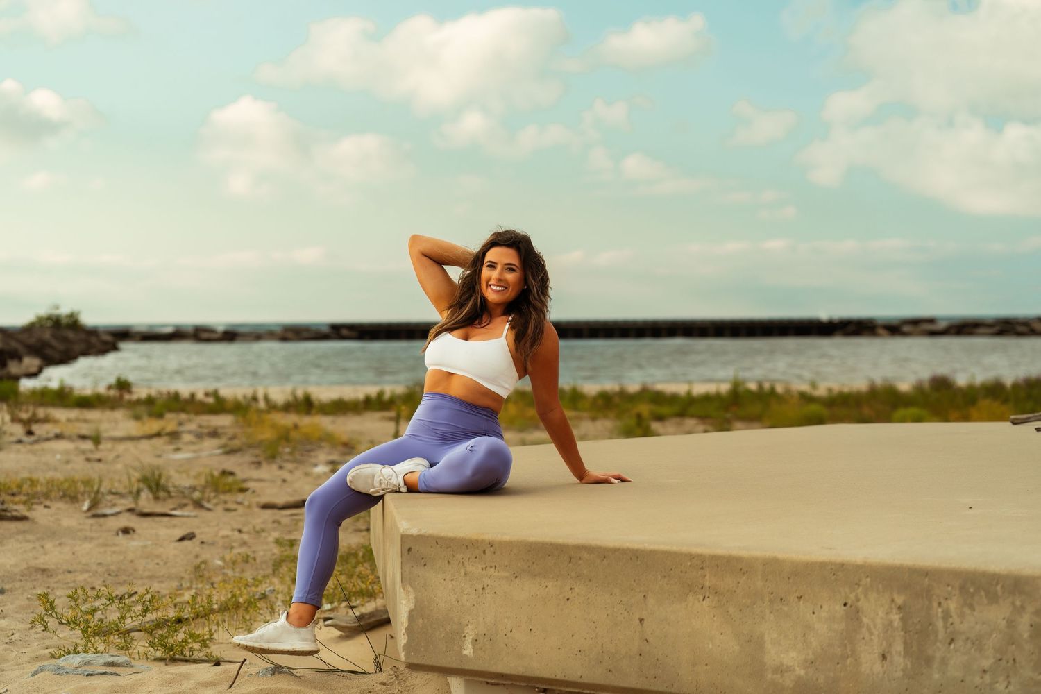 Woman in workout attire poses on concrete ledge near coastal waterfront.