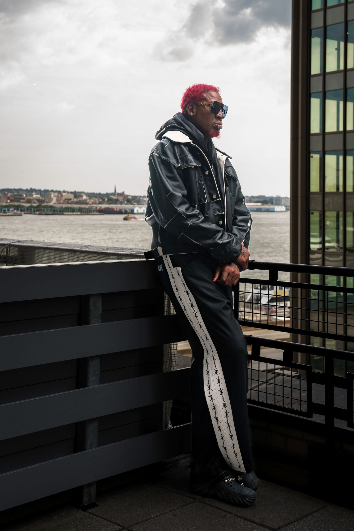 Person in dark jacket sits contemplatively on balcony overlooking cityscape.