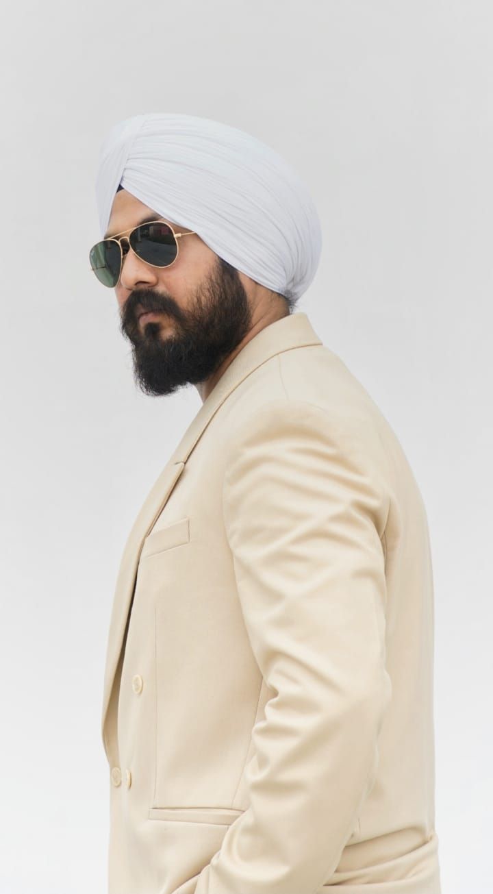 Person in cream blazer and white turban with sunglasses against white backdrop.