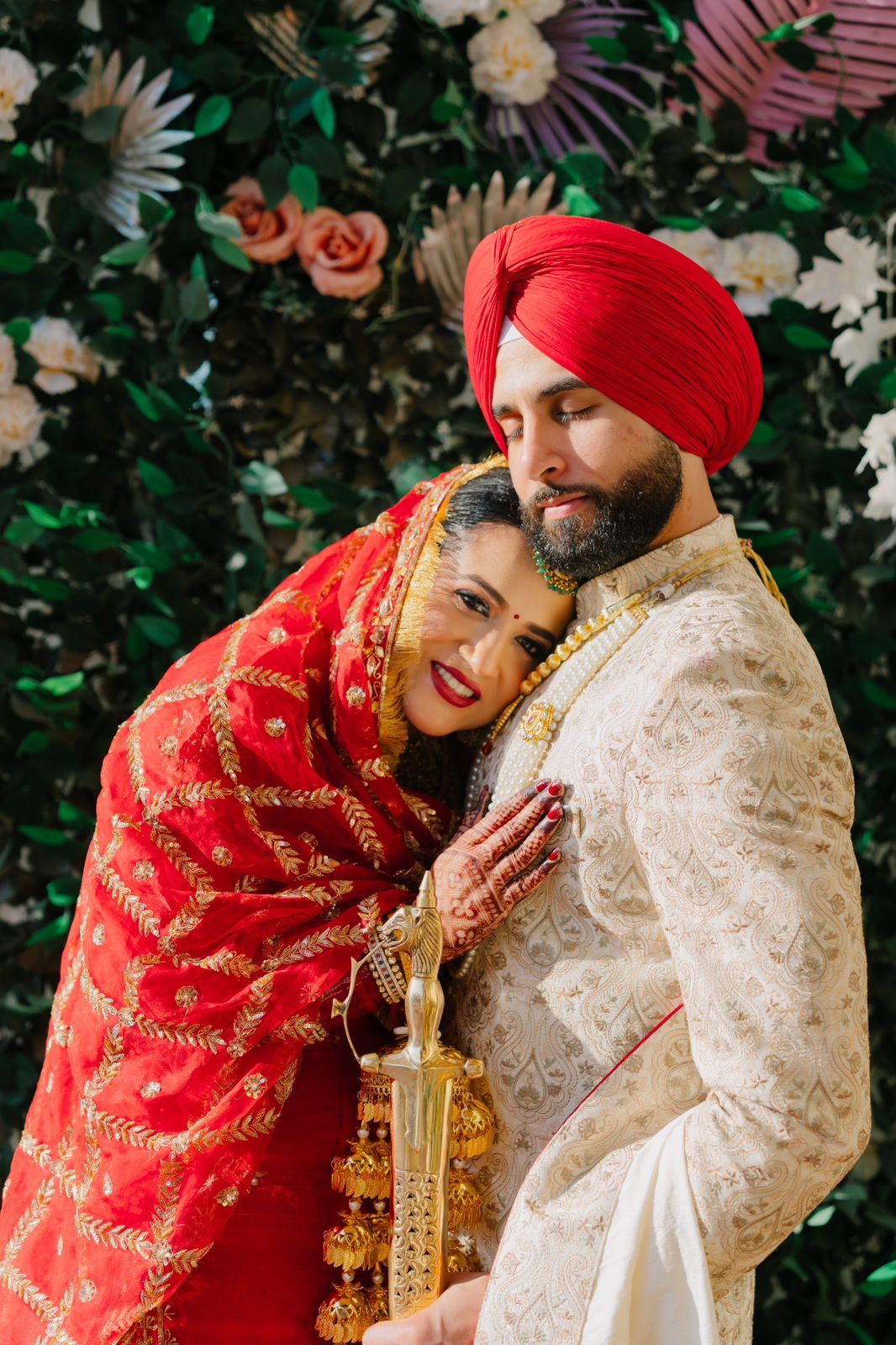 A couple in traditional Sikh wedding attire embraces lovingly against a floral backdrop.