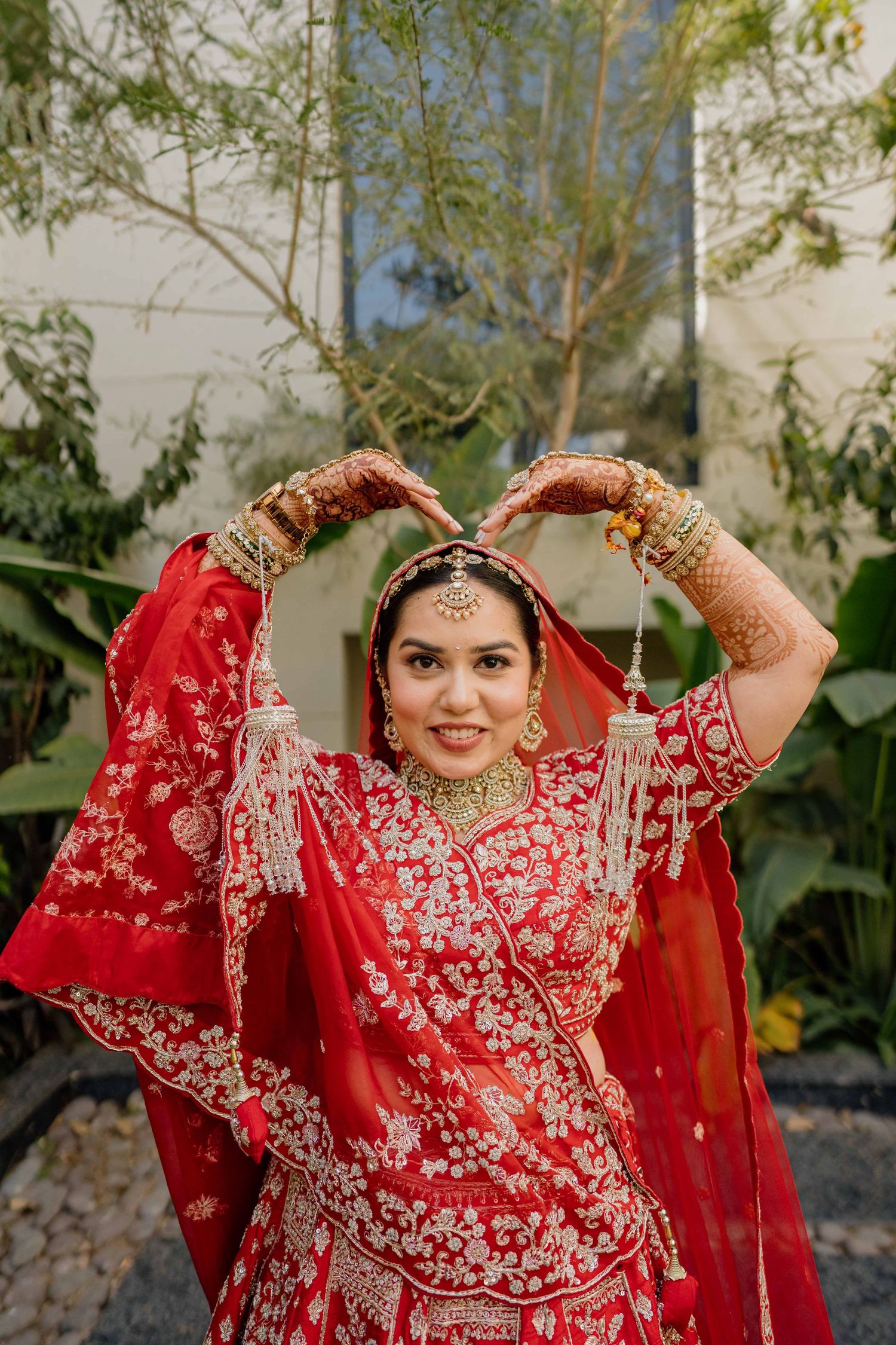 Bride in stunning red embroidered lehenga poses playfully against natural backdrop.