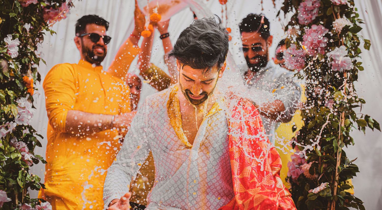 Haldi celebration with people throwing vibrant orange and pink powder.