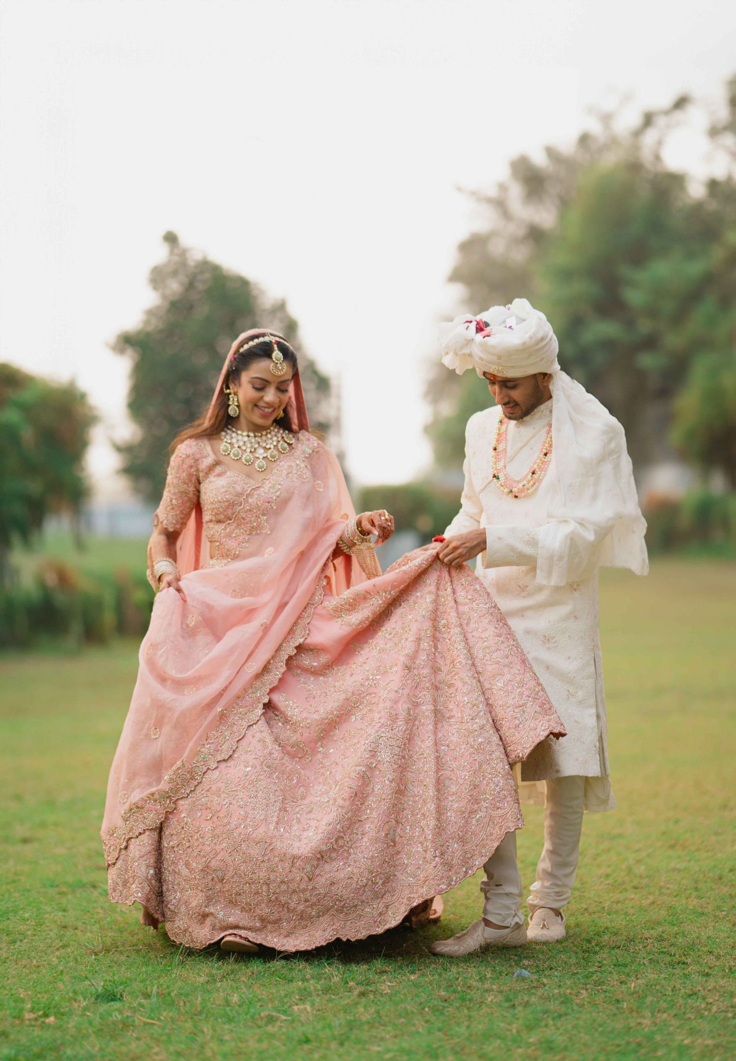 A couple in traditional Indian wedding attire stands on grass, with the bride wearing a pink embellished lehenga.