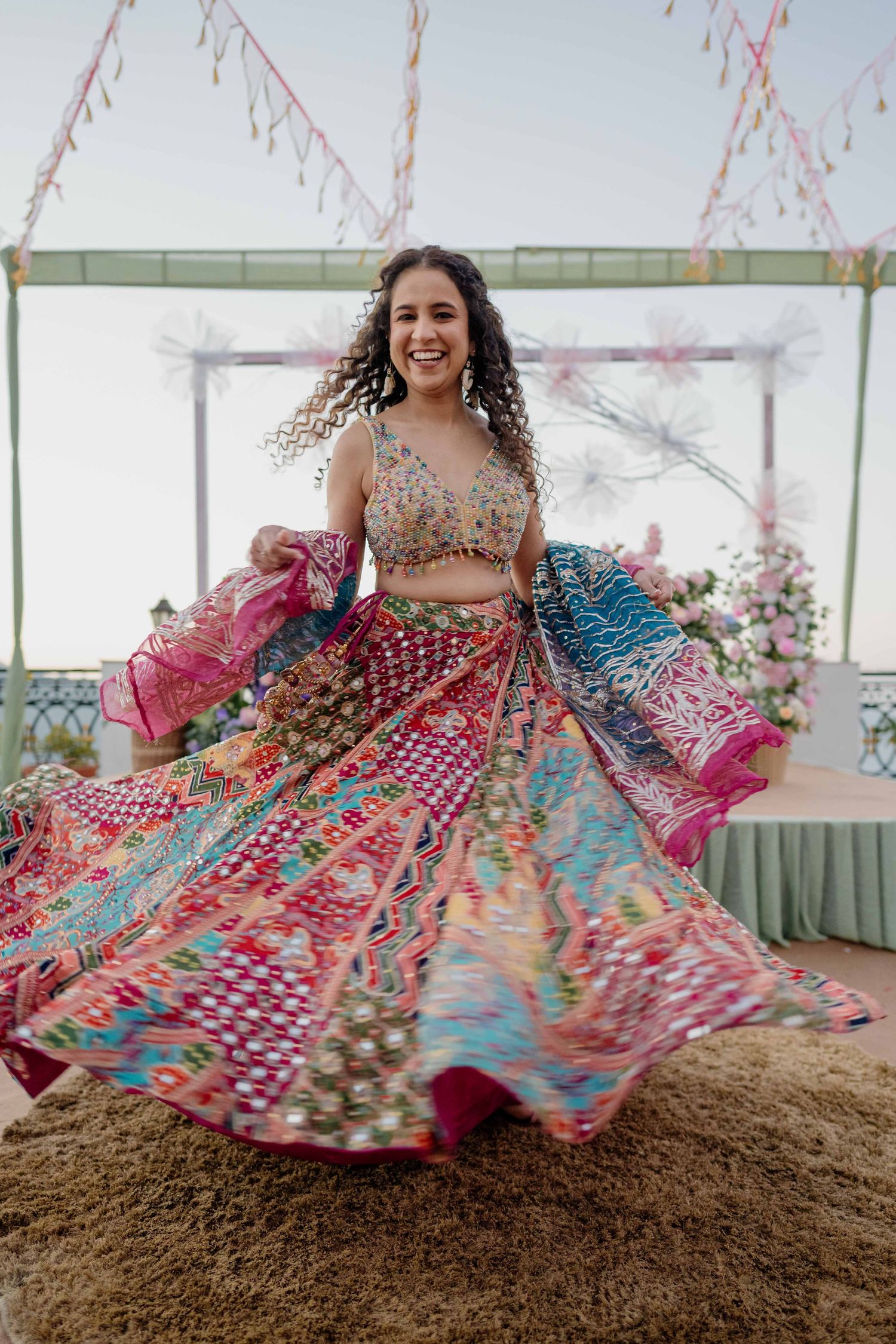 A person in a colorful lehenga twirls on dirt ground, creating a vibrant display of pink and blue fabric in motion.