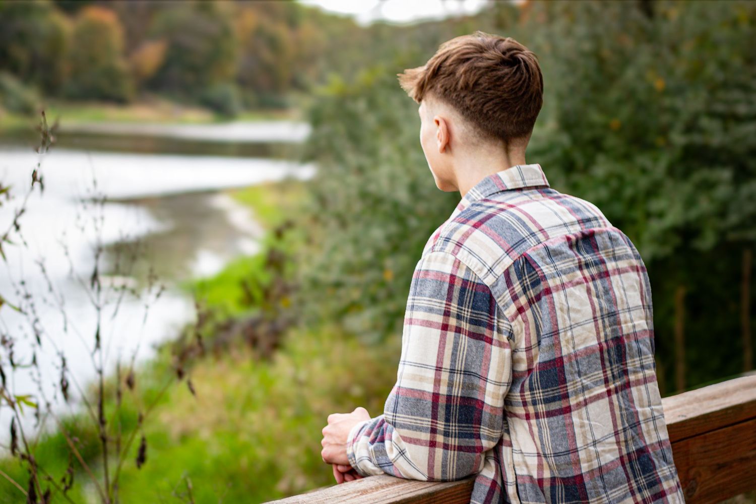 Someone in a plaid shirt sits alone on a wooden railing overlooking a peaceful lake surrounded by autumn foliage.