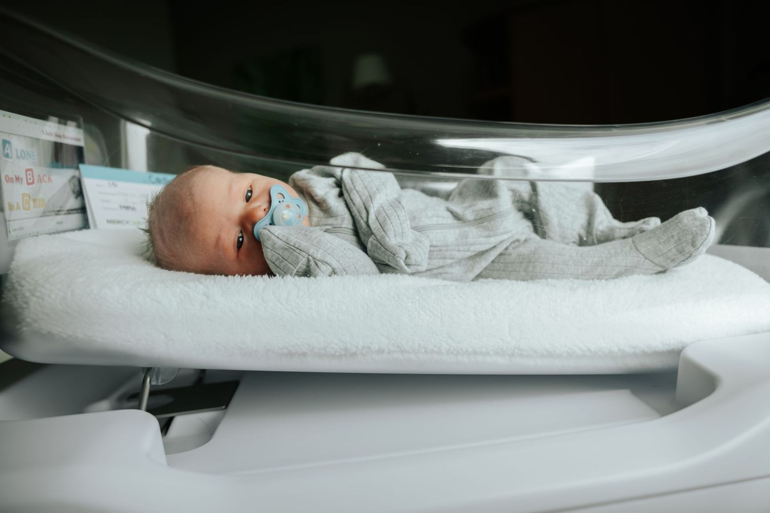 Newborn baby with pacifier lying in a modern hospital bassinet, dressed in gray onesie.