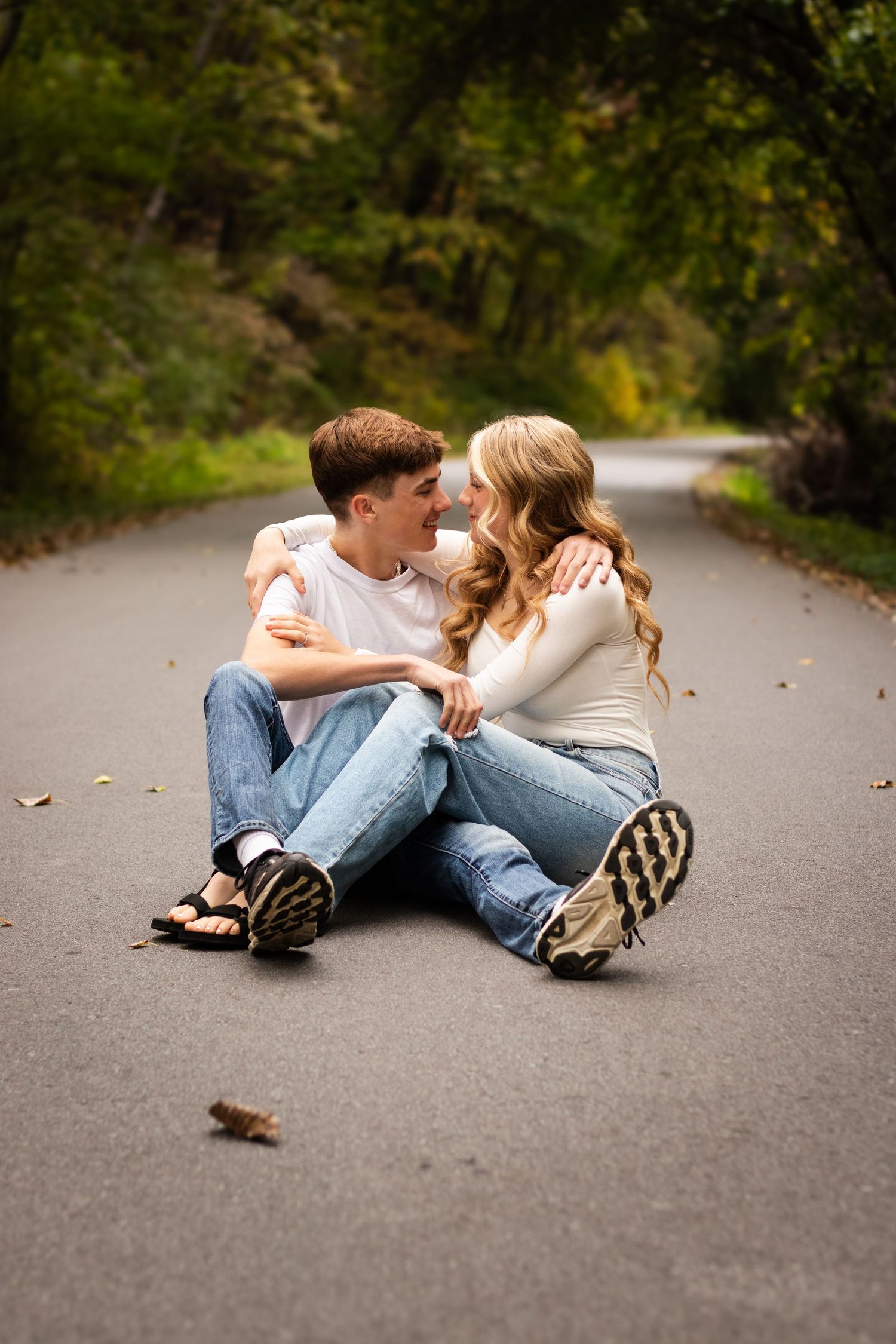 A couple sits intimately together on a winding forest road surrounded by green trees on an autumn day.