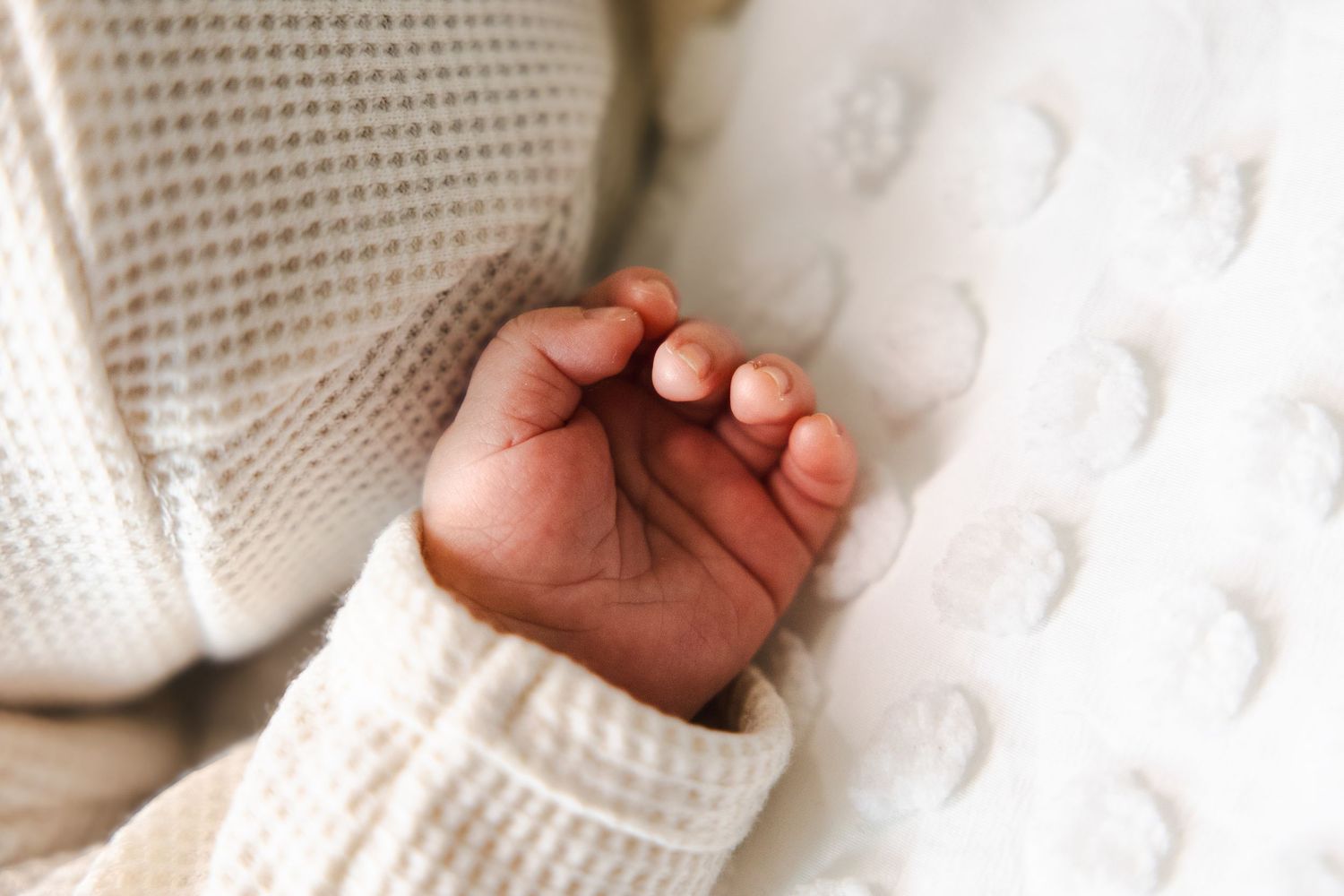 Close-up of a newborn baby's tiny hand resting on a soft white textured blanket, wearing a cream knit sweater.