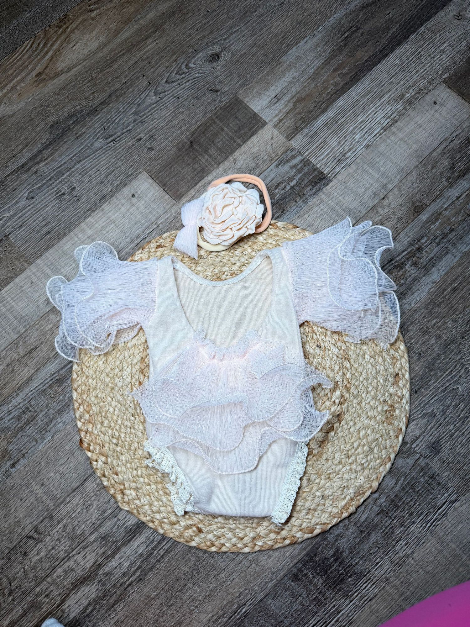 White ruffled baby romper with floral headband displayed on a woven straw mat on wood floor.