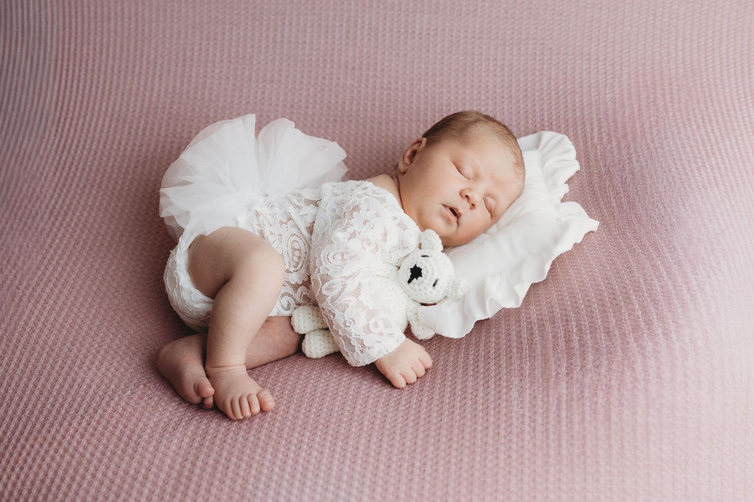 Newborn baby sleeping peacefully in white lace outfit cuddling a soft teddy bear on a pink blanket.