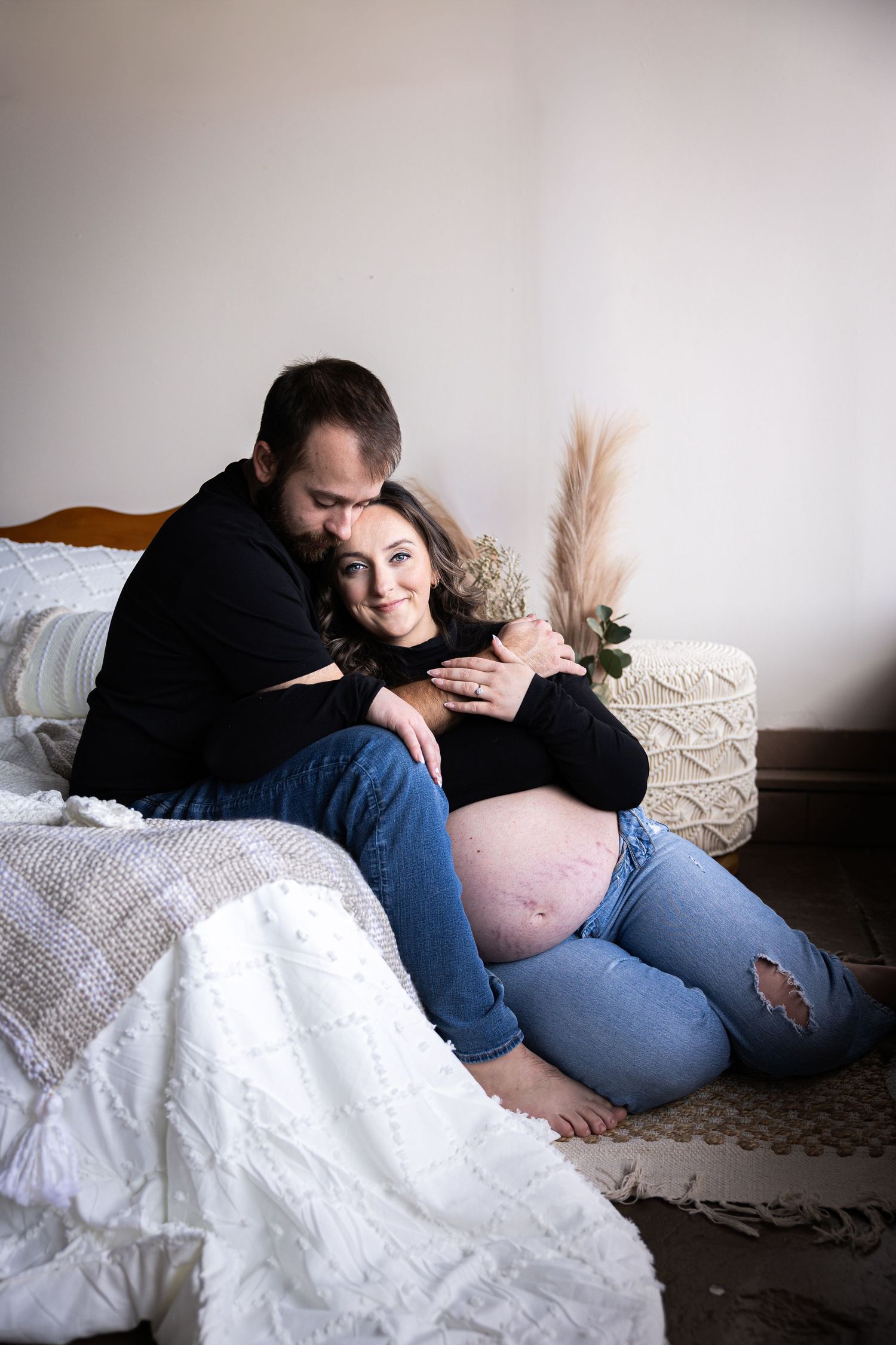 Tender maternity moment photographed on white bedding with soft natural lighting.