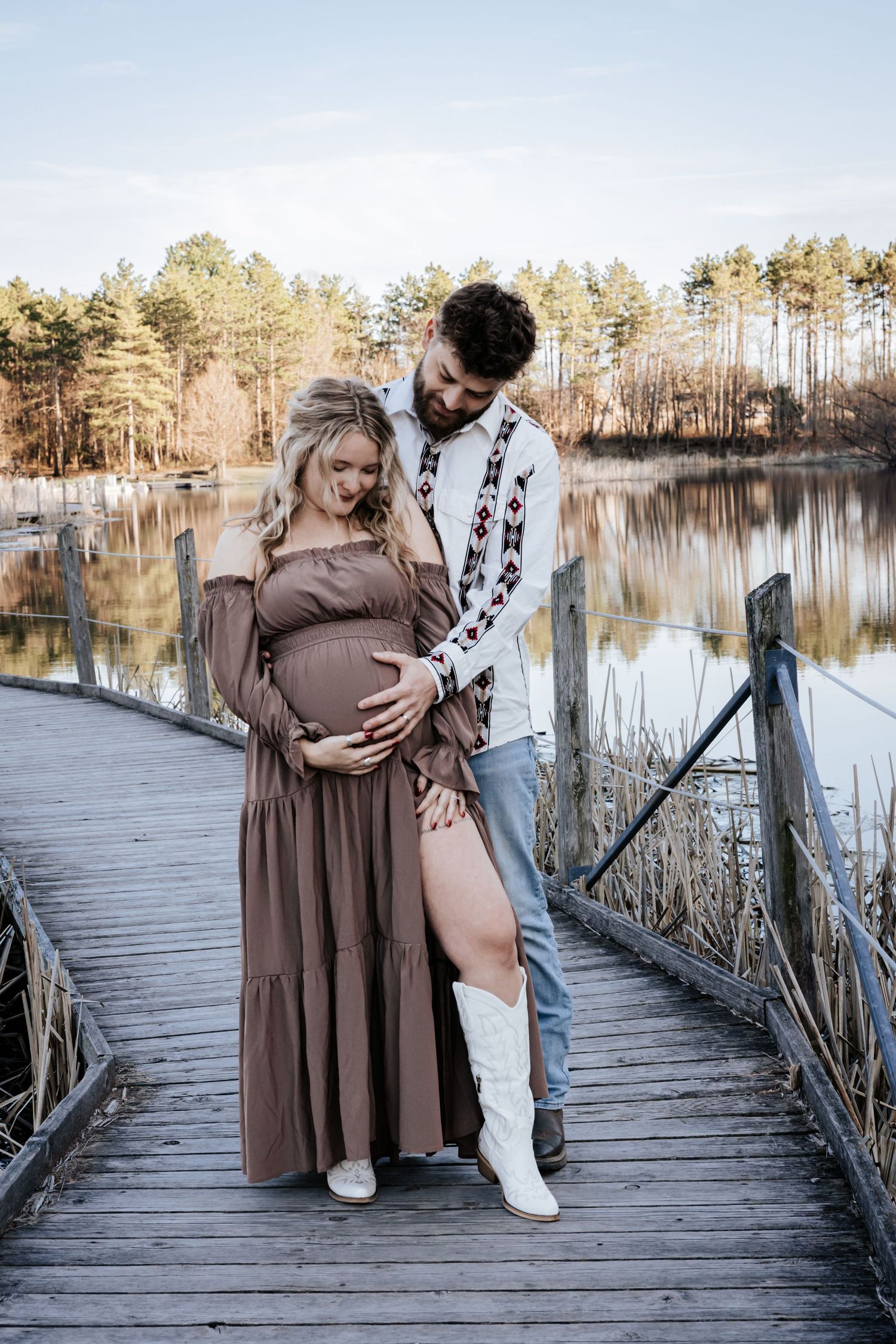 Pregnant couple standing on a wooden dock by a lake, woman in brown maxi dress, man in embroidered shirt, embracing.