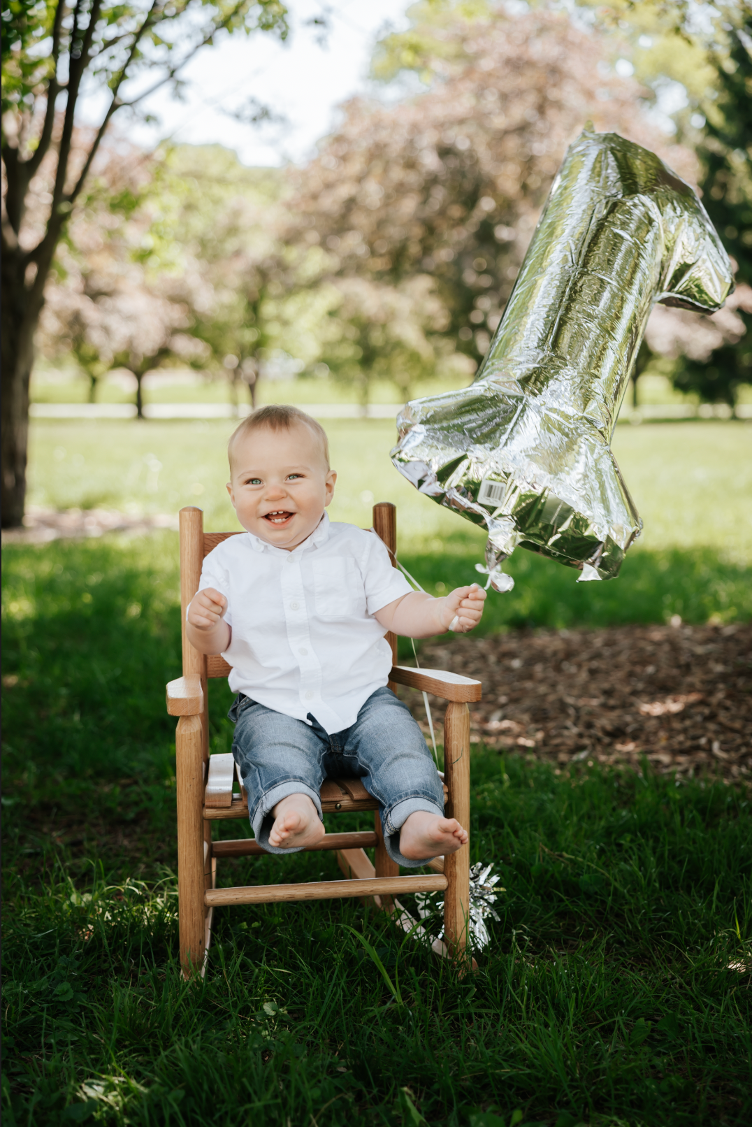 Happy baby boy sitting in wooden chair holding silver number 1 balloon in a park on his first birthday.