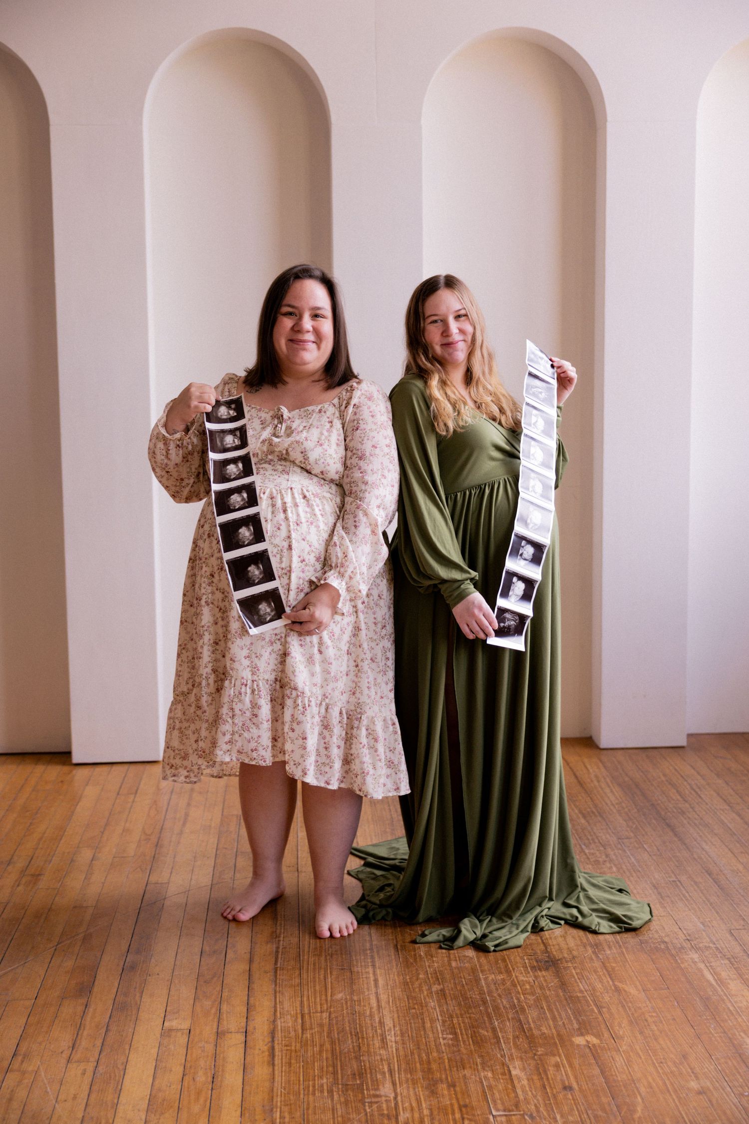Two people in flowing dresses sit on wooden floor holding ultrasound photos while sharing a joyful pregnancy announcement moment.