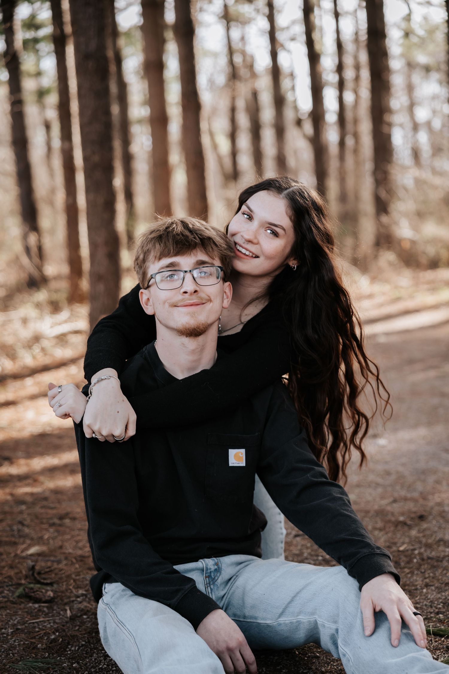 A young couple poses in a pine forest, the girl hugging the boy from behind as he sits on the ground smiling.