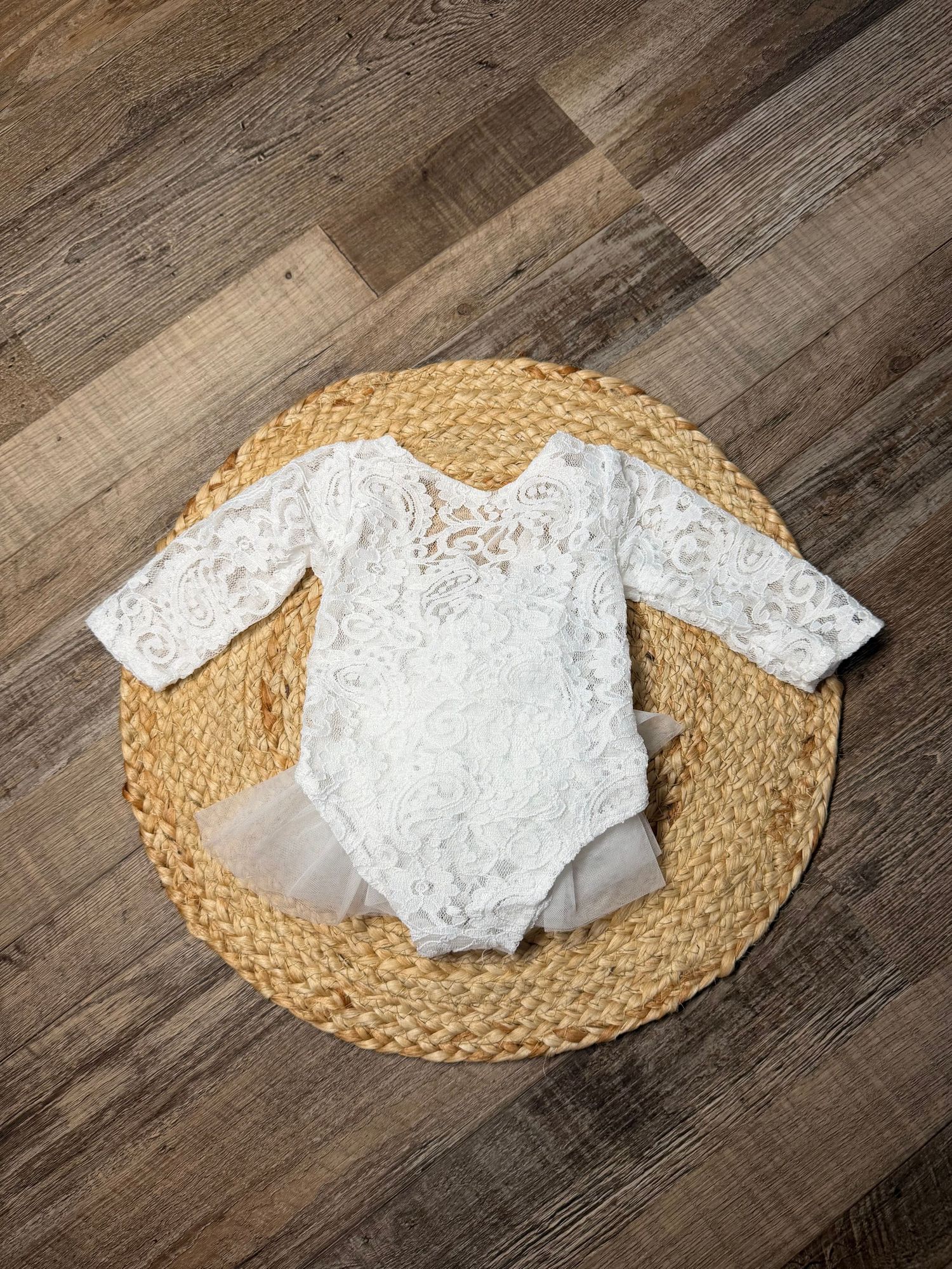 White lace long-sleeve baby bodysuit with tutu skirt displayed on woven mat on wood floor.