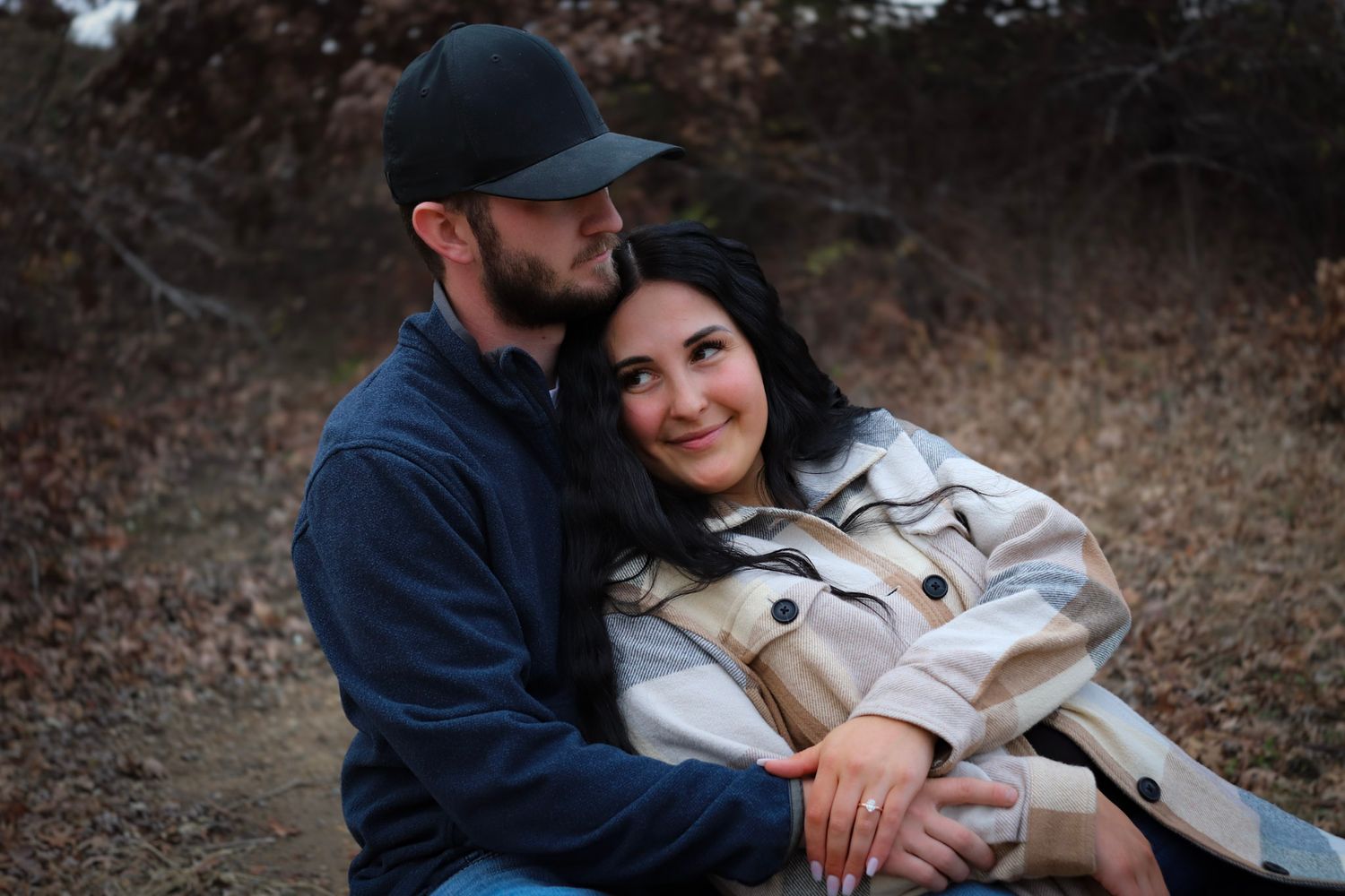 A couple embraces outdoors on a fall evening, with one wearing a navy jacket and baseball cap and the other in a beige coat.