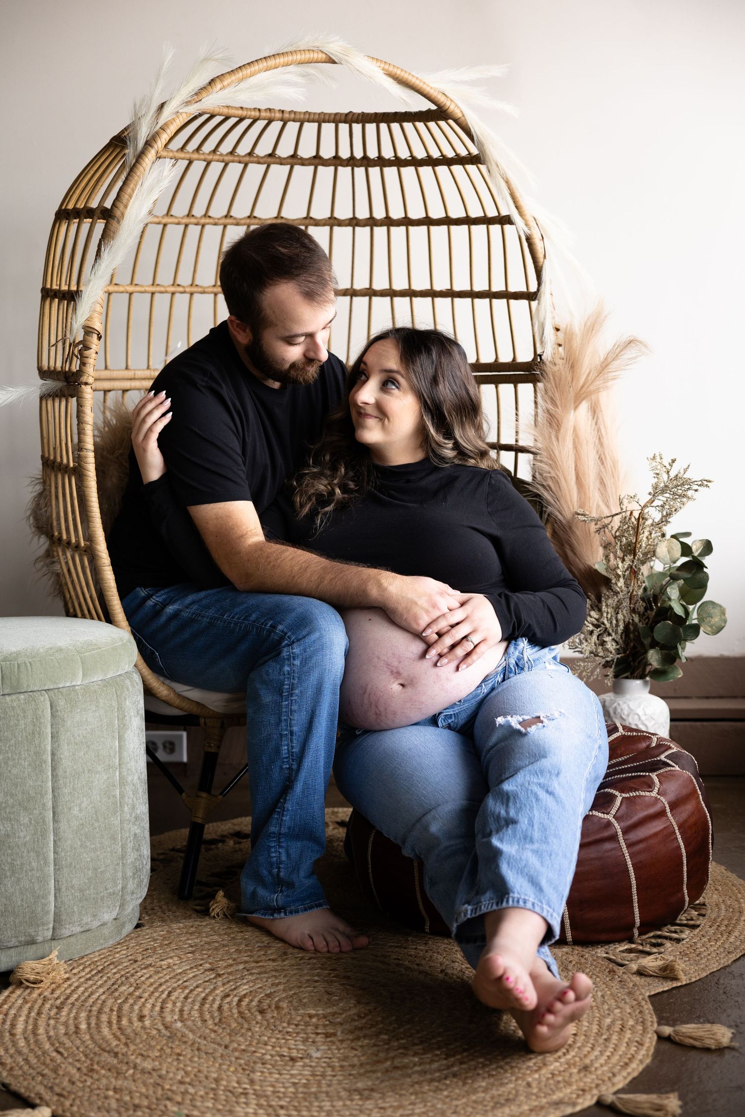 Expectant couple relaxing in rattan chair with boho decor and jute rug.
