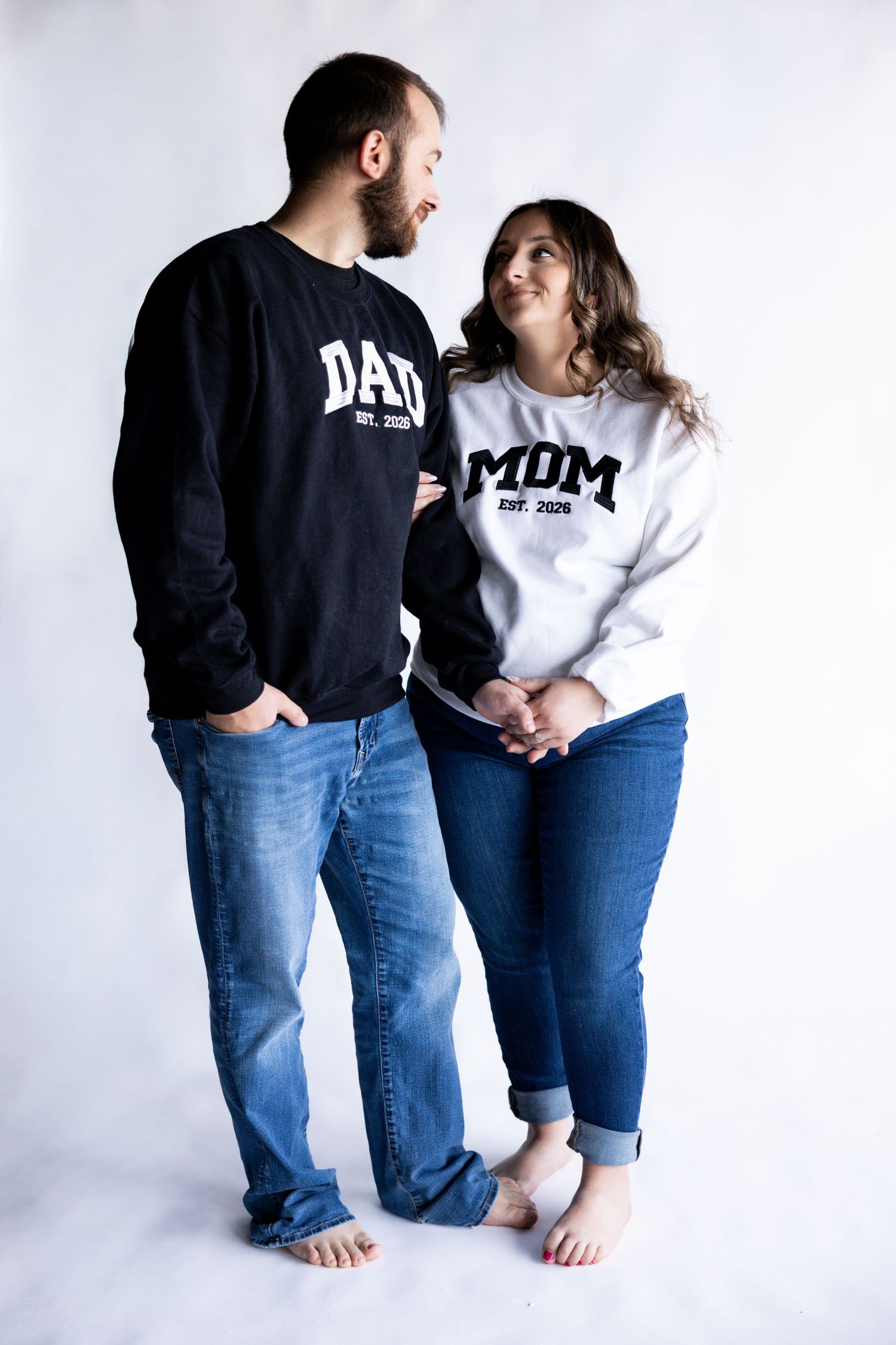 Couple wearing matching Mom and Dad sweatshirts pose barefoot against white background.