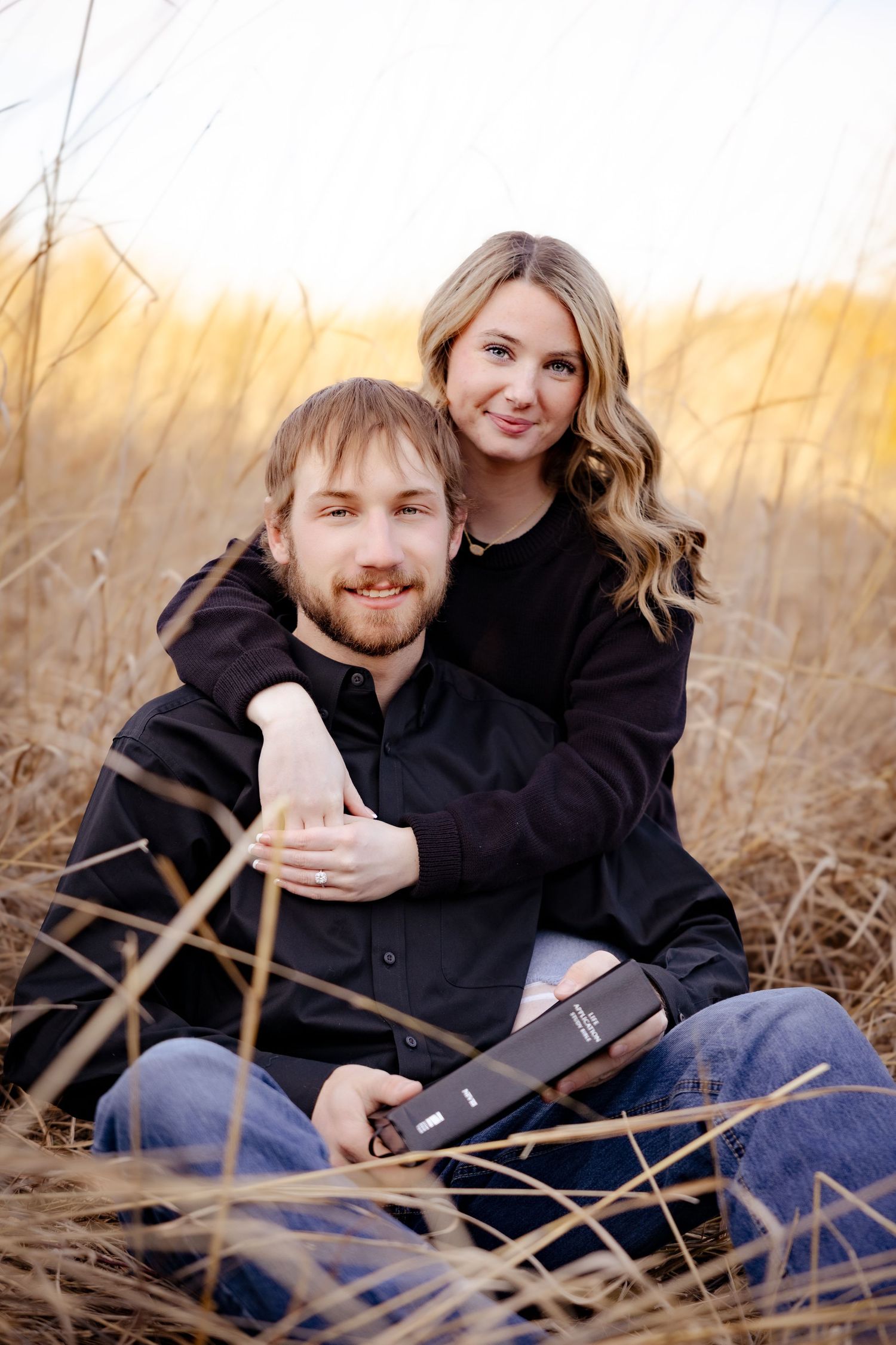 Couple sitting in golden field, woman embracing man from behind, both in black shirts, holding a Bible at sunset.