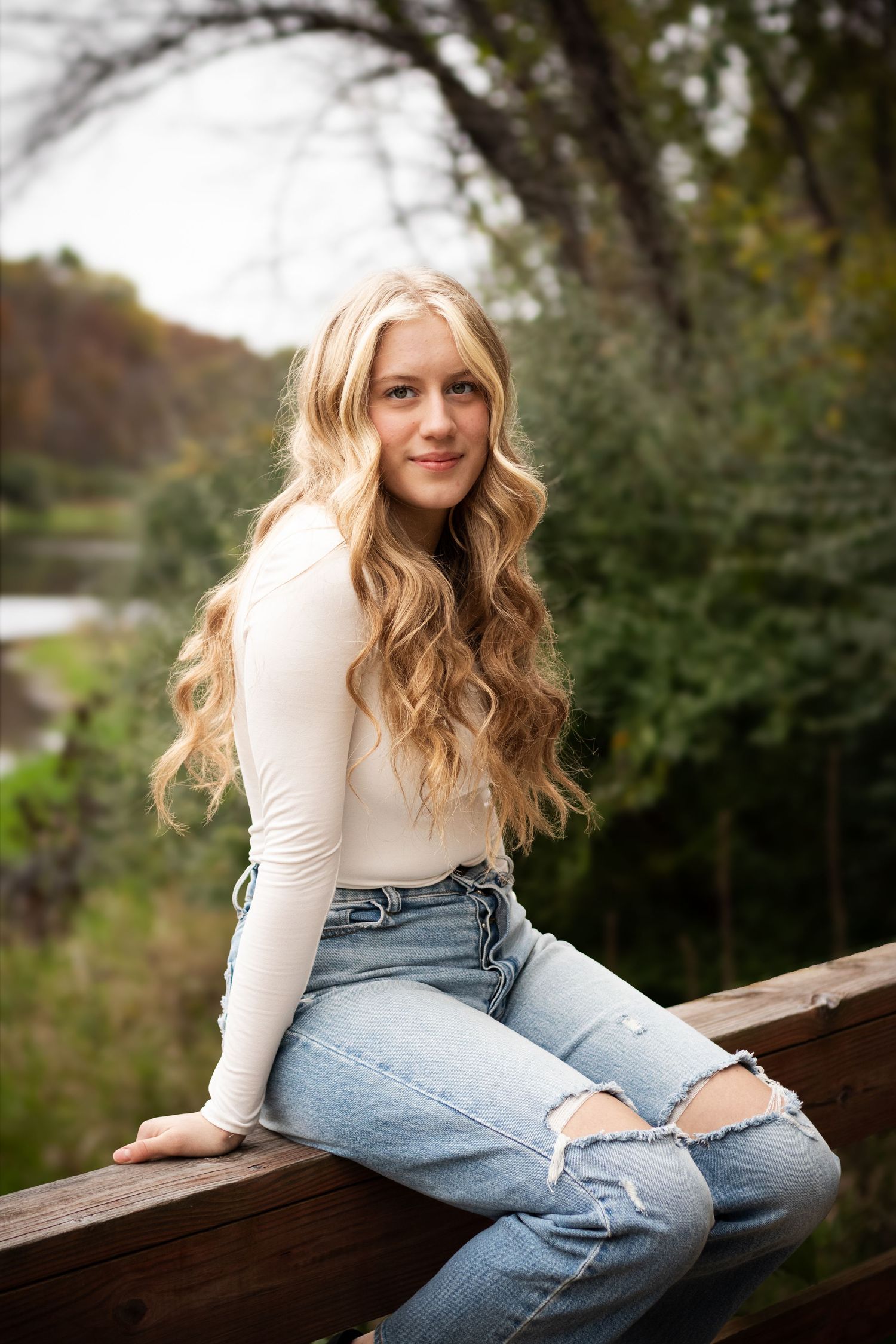 A person in a cream sweater and distressed jeans poses casually on a wooden bench in an outdoor setting.