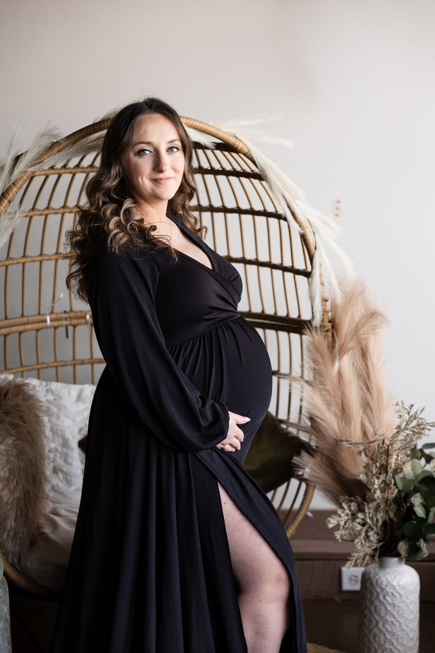 Maternity photoshoot featuring a black dress against a rattan peacock chair with dried pampas grass decor.