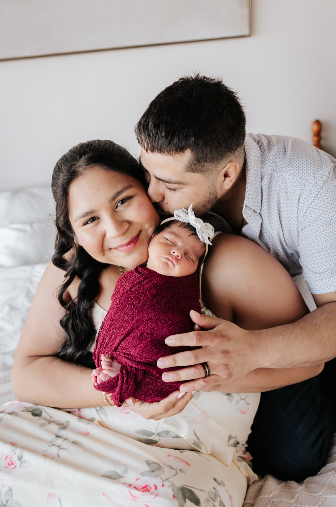 A couple sits on a bed, smiling as the man kisses the woman's cheek while she holds their newborn baby wrapped in red.