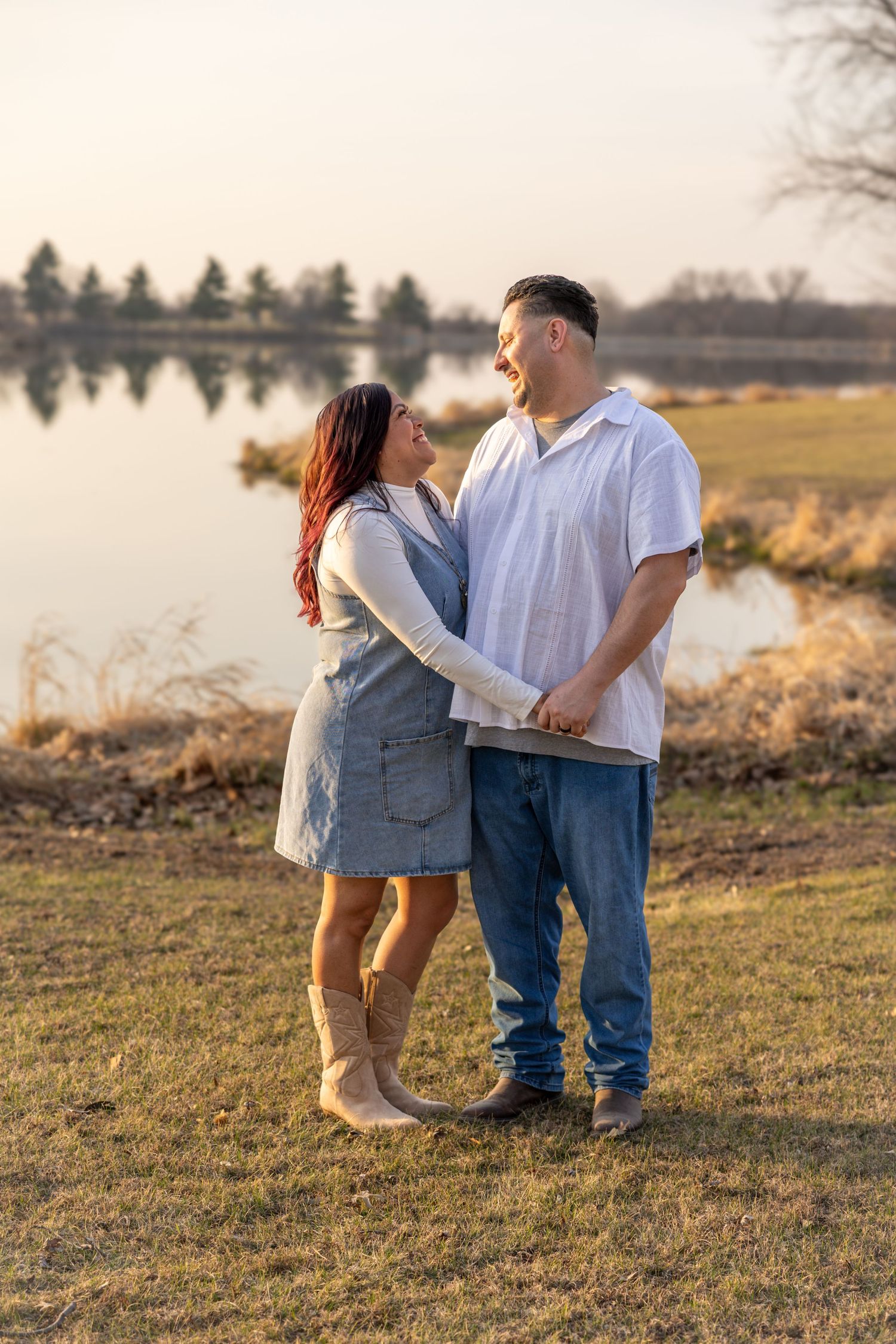A couple shares a romantic moment in a golden field at sunset by a lake.