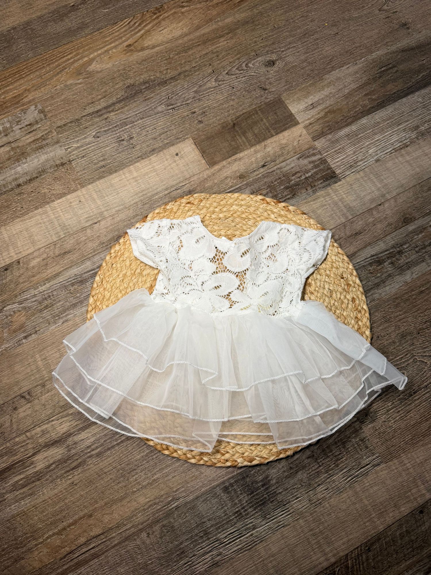 White lace and tulle baby girl dress with ruffled skirt displayed on woven mat over wood floor.