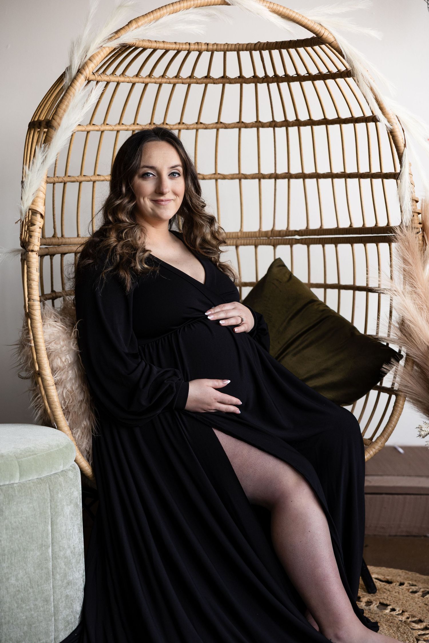 A maternity portrait in a rattan chair showing an elegant black dress and bohemian styling.