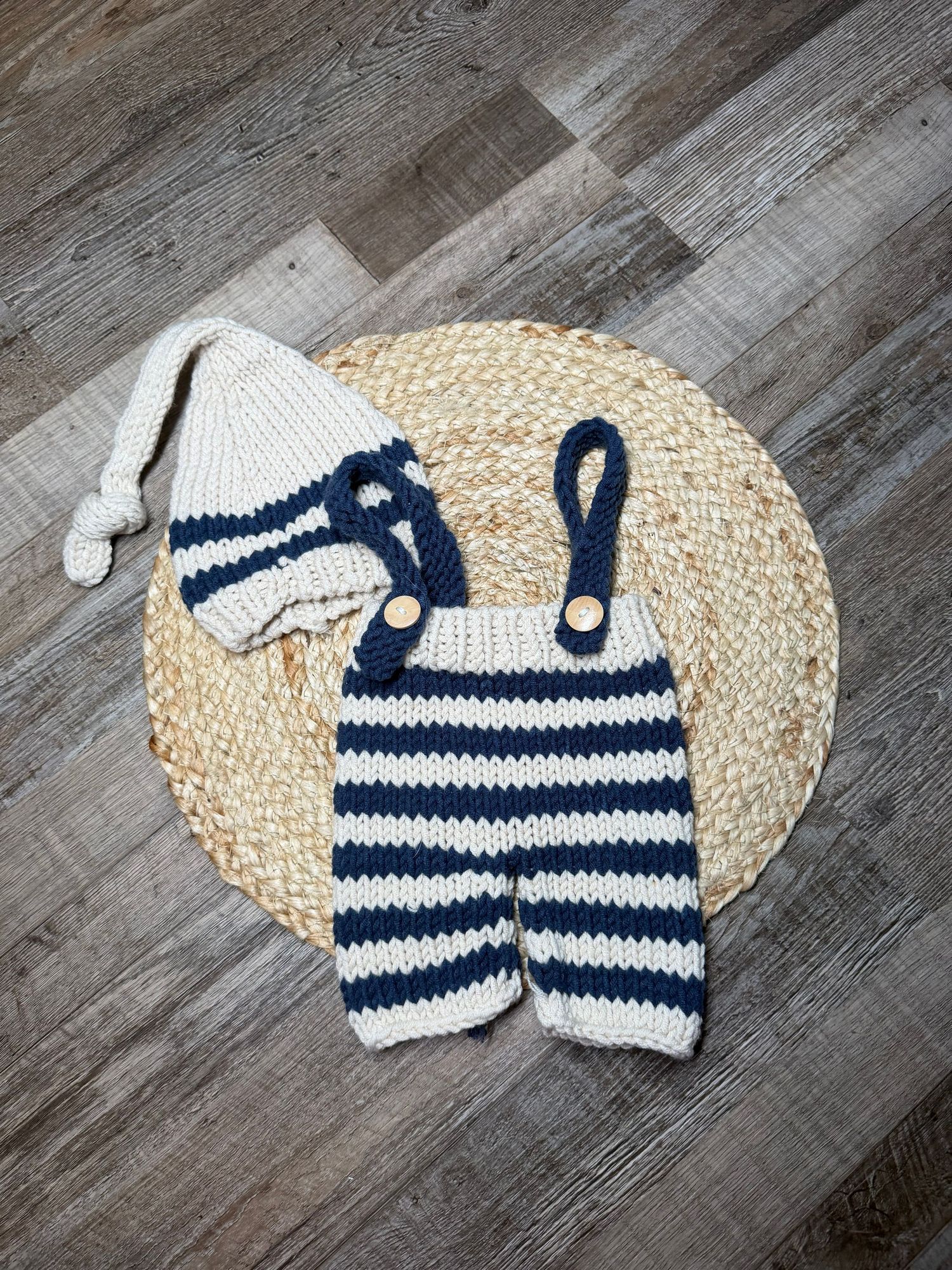 Hand-knitted navy and white striped newborn suspender pants and matching elf hat set displayed on wicker mat.