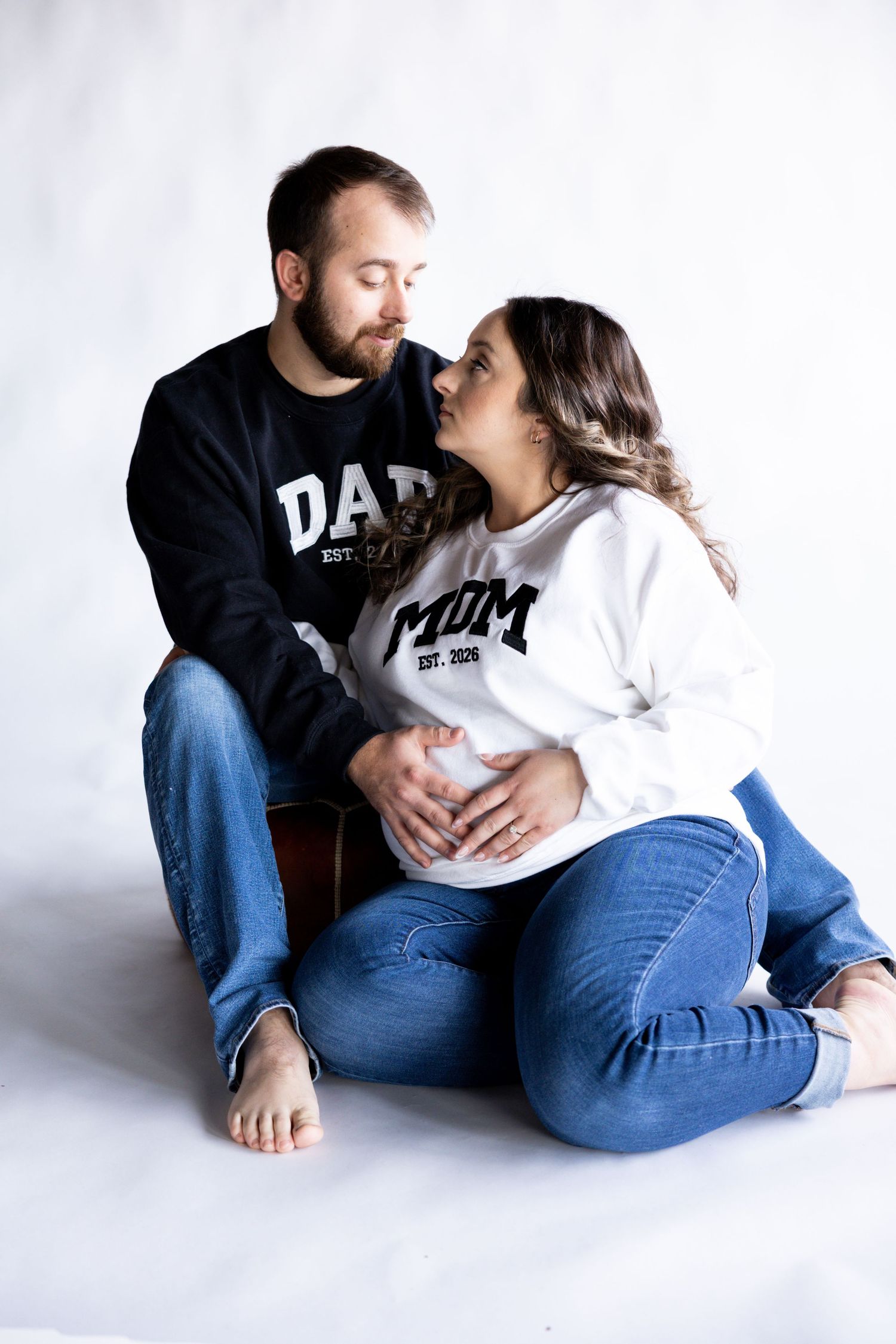 A couple wearing matching Dad and Mom shirts embrace while sitting on a white floor in a romantic photo series.