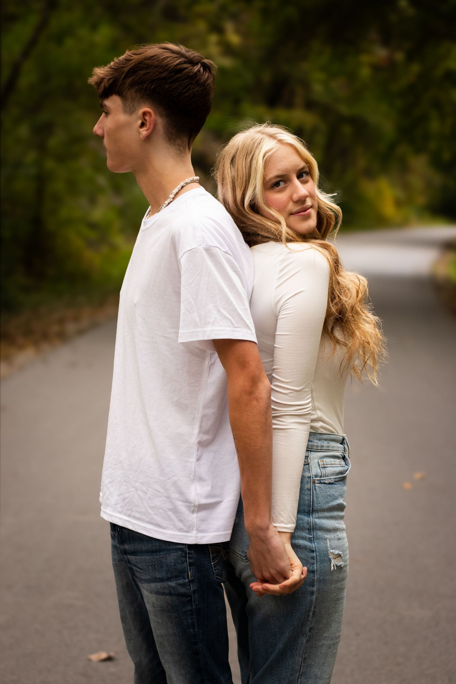A couple in casual attire stands together on a paved road surrounded by greenery in a romantic pose.