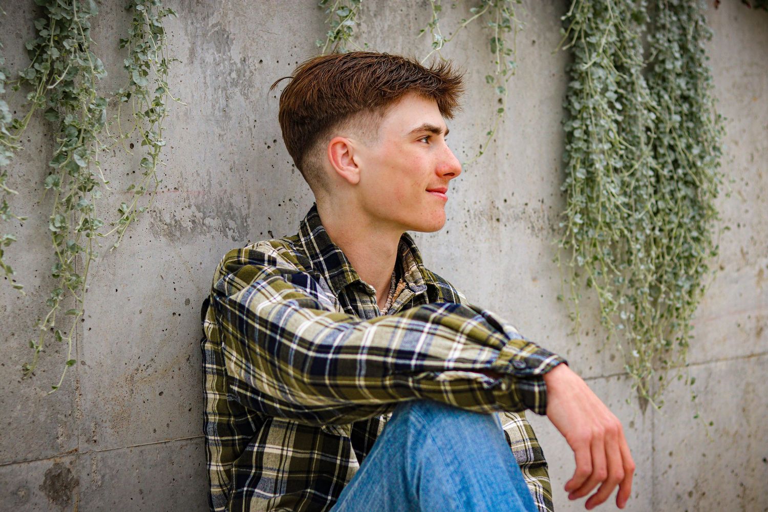 A person in a plaid shirt and blue jeans sits against a concrete wall with hanging plants.