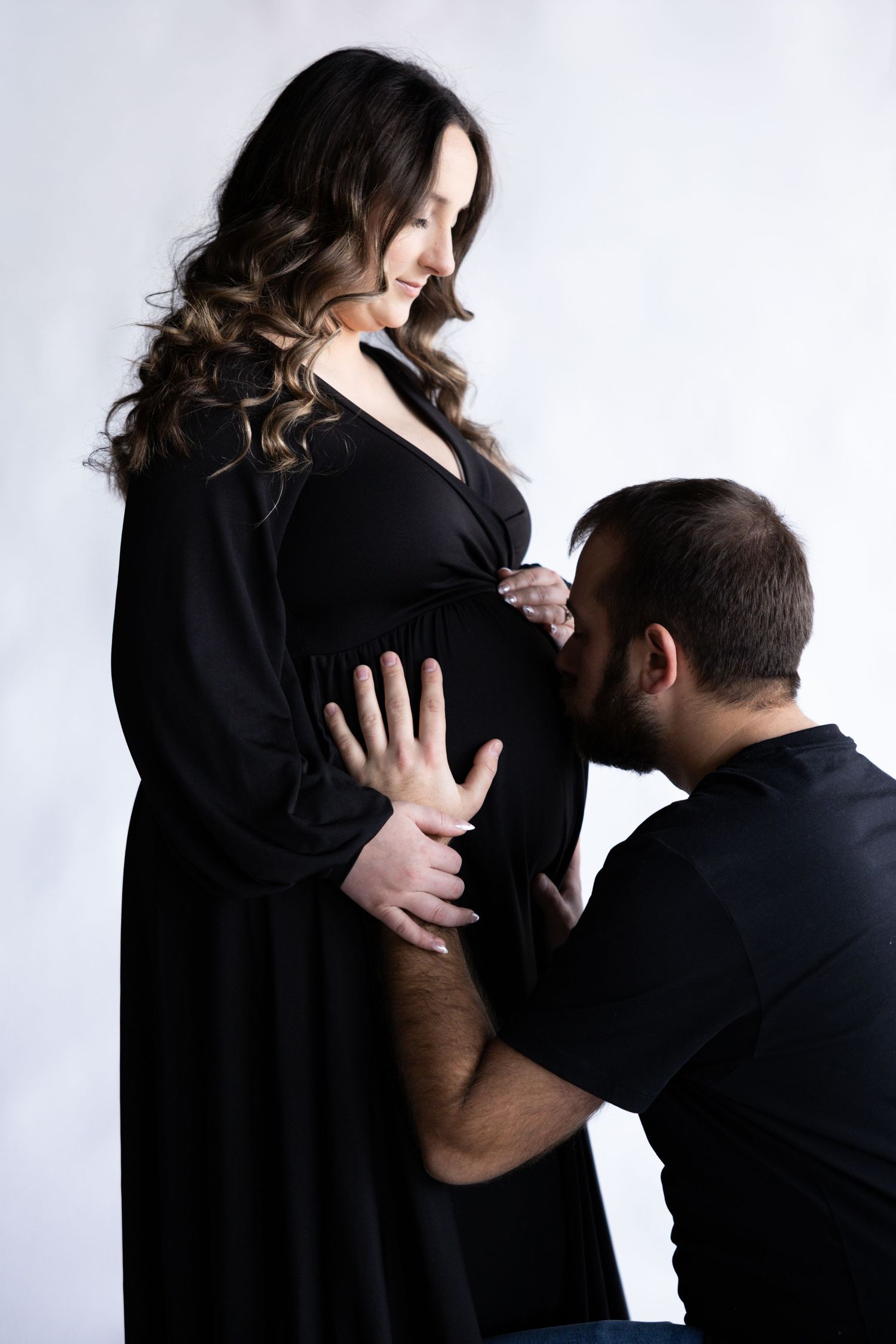 Intimate maternity photo session featuring couple in black outfits against white backdrop.