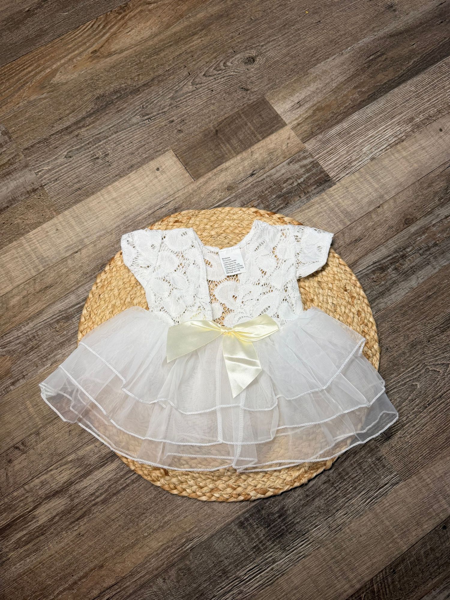 White lace short-sleeve baby dress with yellow bow and tiered tulle skirt on woven mat.