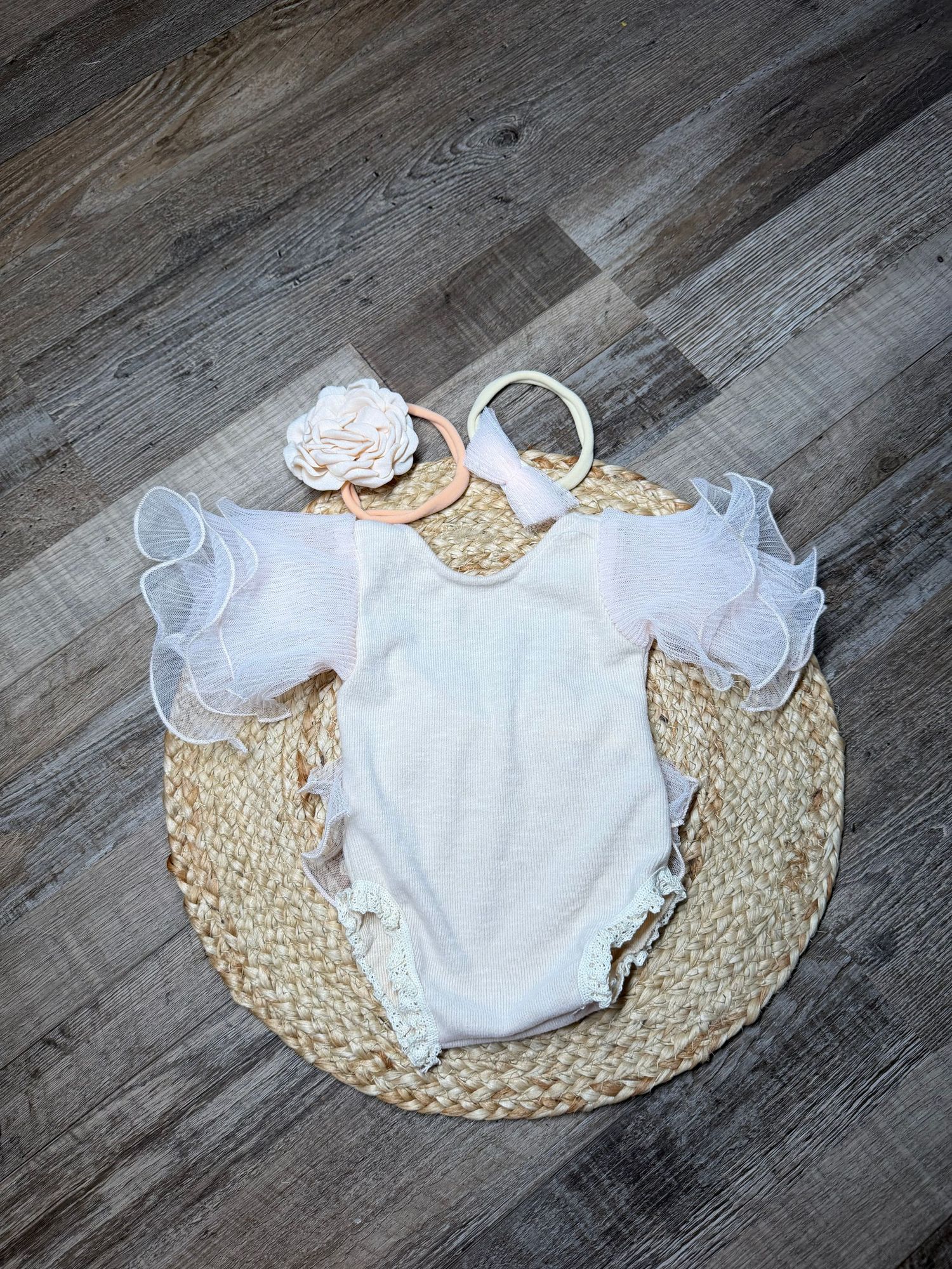 White ruffled sleeve baby romper with lace trim and floral headband displayed on woven mat on wood floor.