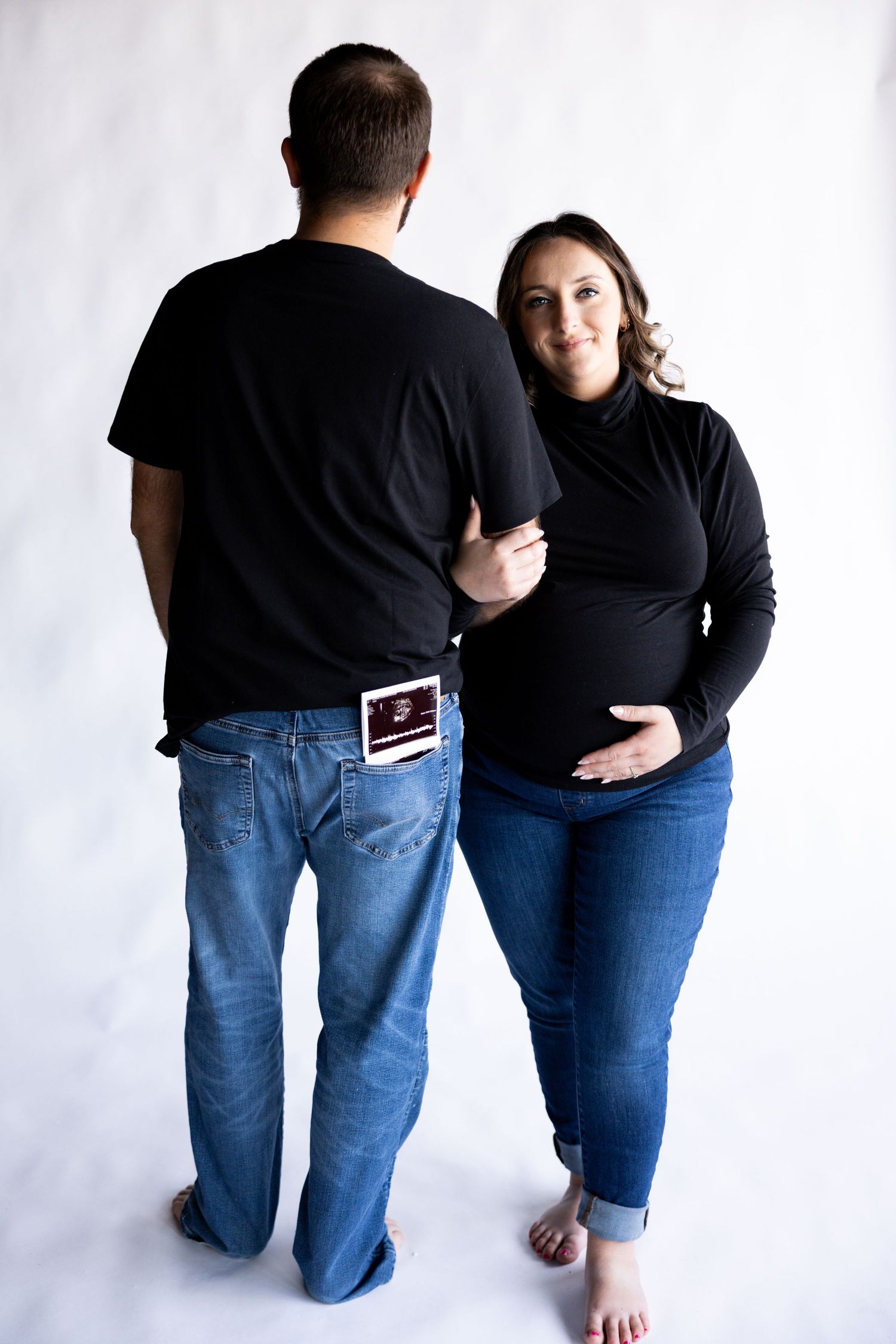 Minimalist maternity photo against white backdrop with ultrasound photo visible in back pocket.