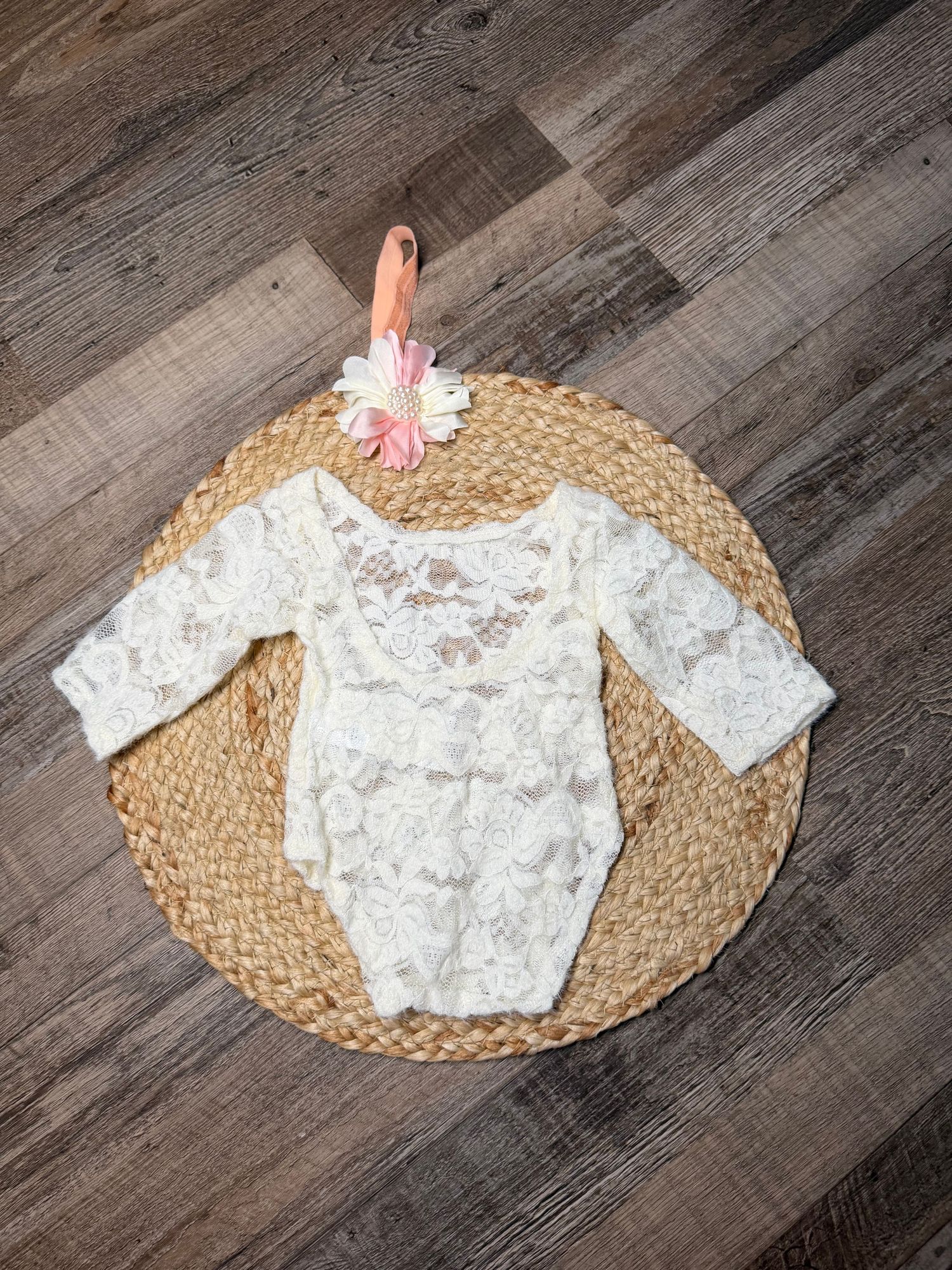 White lace long-sleeve baby romper with floral headband displayed on woven mat on wood floor.