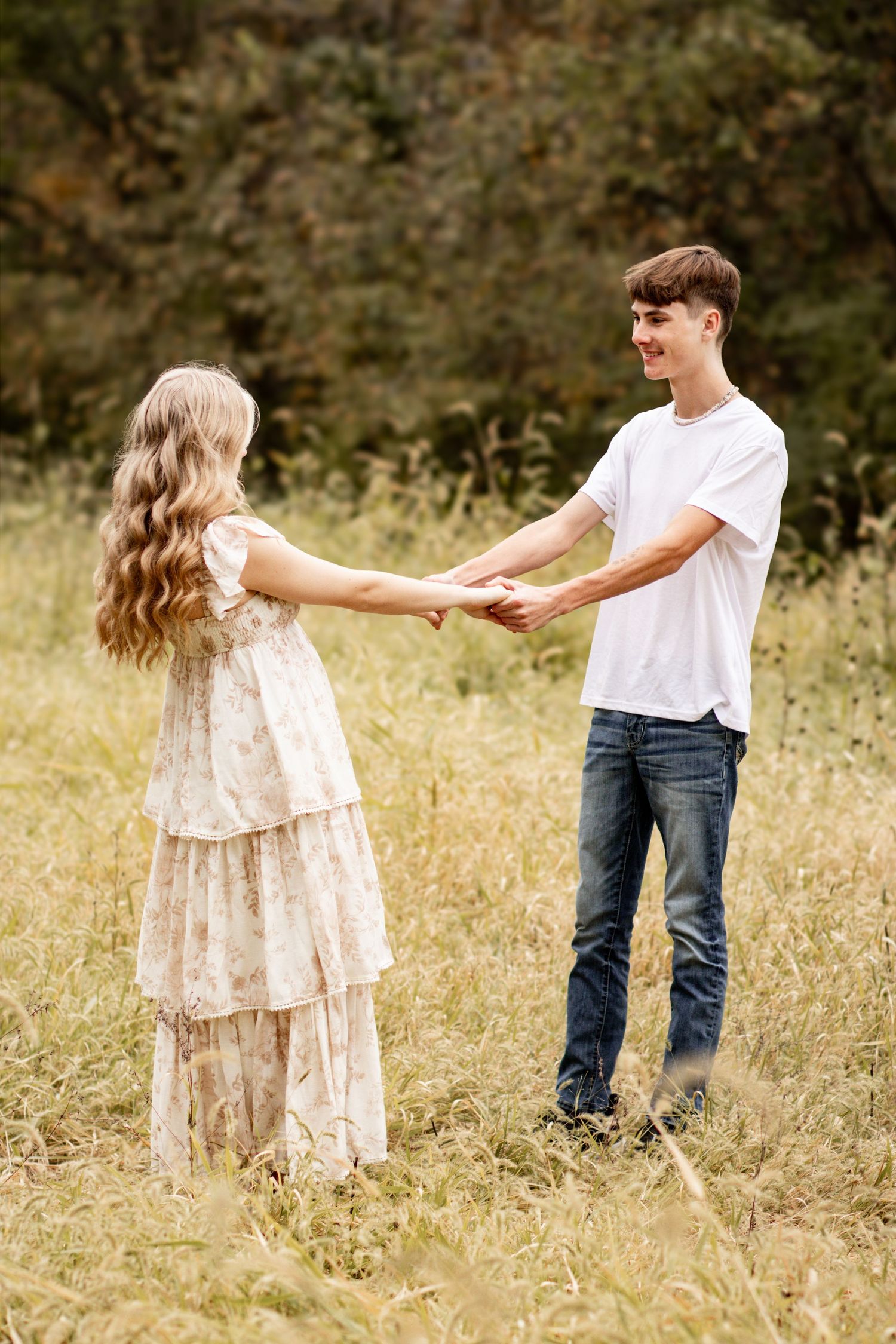 A couple holds hands while standing in a sunlit meadow filled with wildflowers and tall grass.