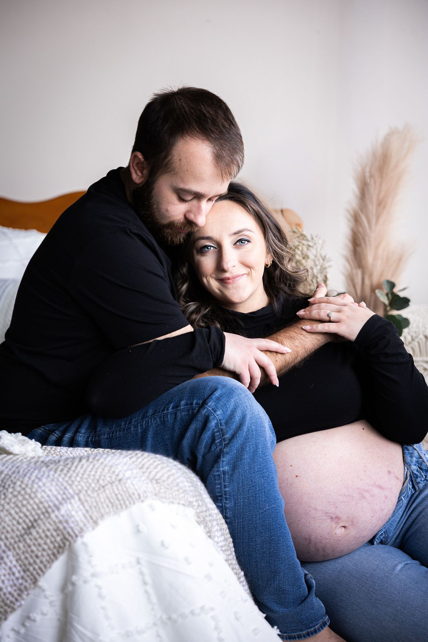 Intimate couple in casual attire resting together on white bedding with soft lighting and pampas grass decor.