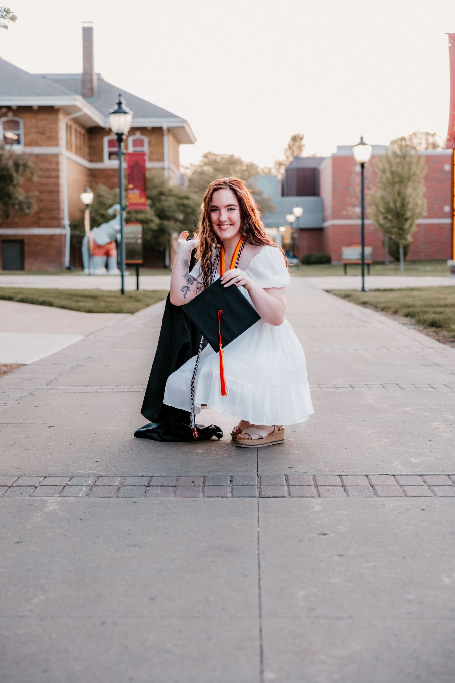 A smiling graduate in a white dress kneels on a campus walkway, holding her black cap with red tassel.