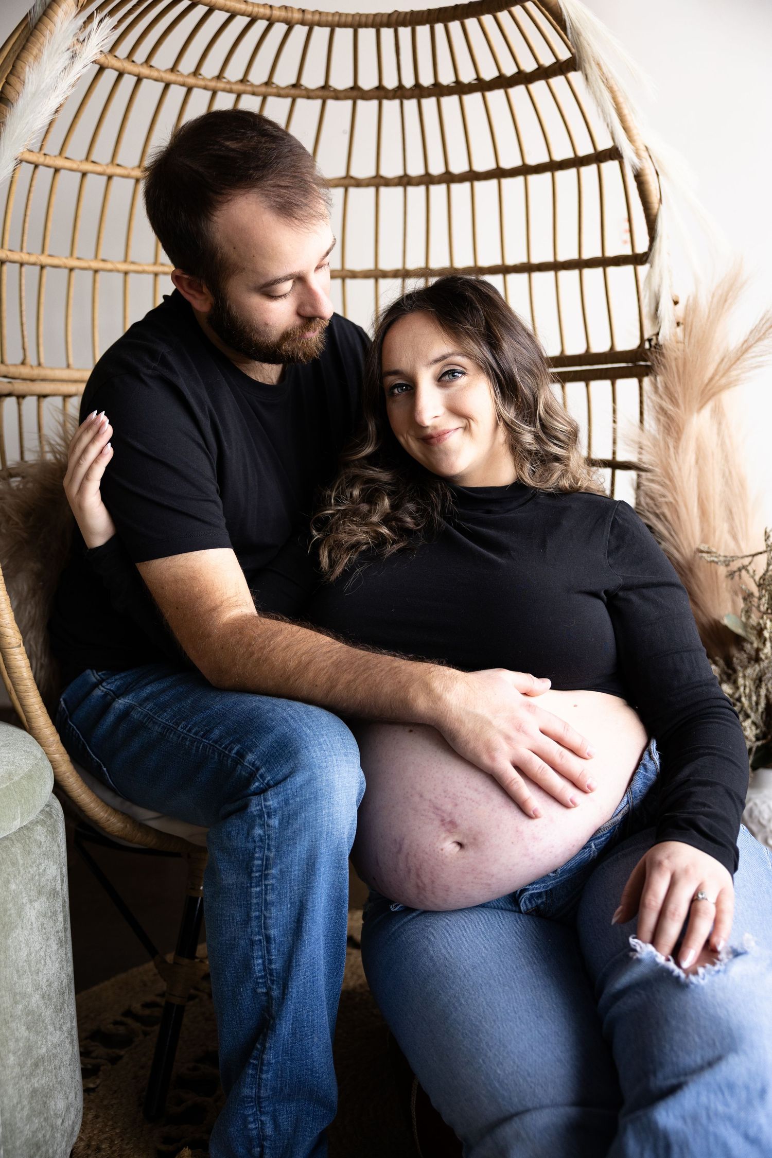 A maternity photoshoot featuring a couple sitting together in a rattan hanging chair with pampas grass decor.