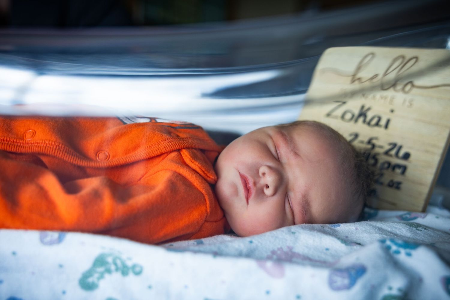 A sleeping baby wrapped in an orange blanket rests peacefully in a hospital bassinet.